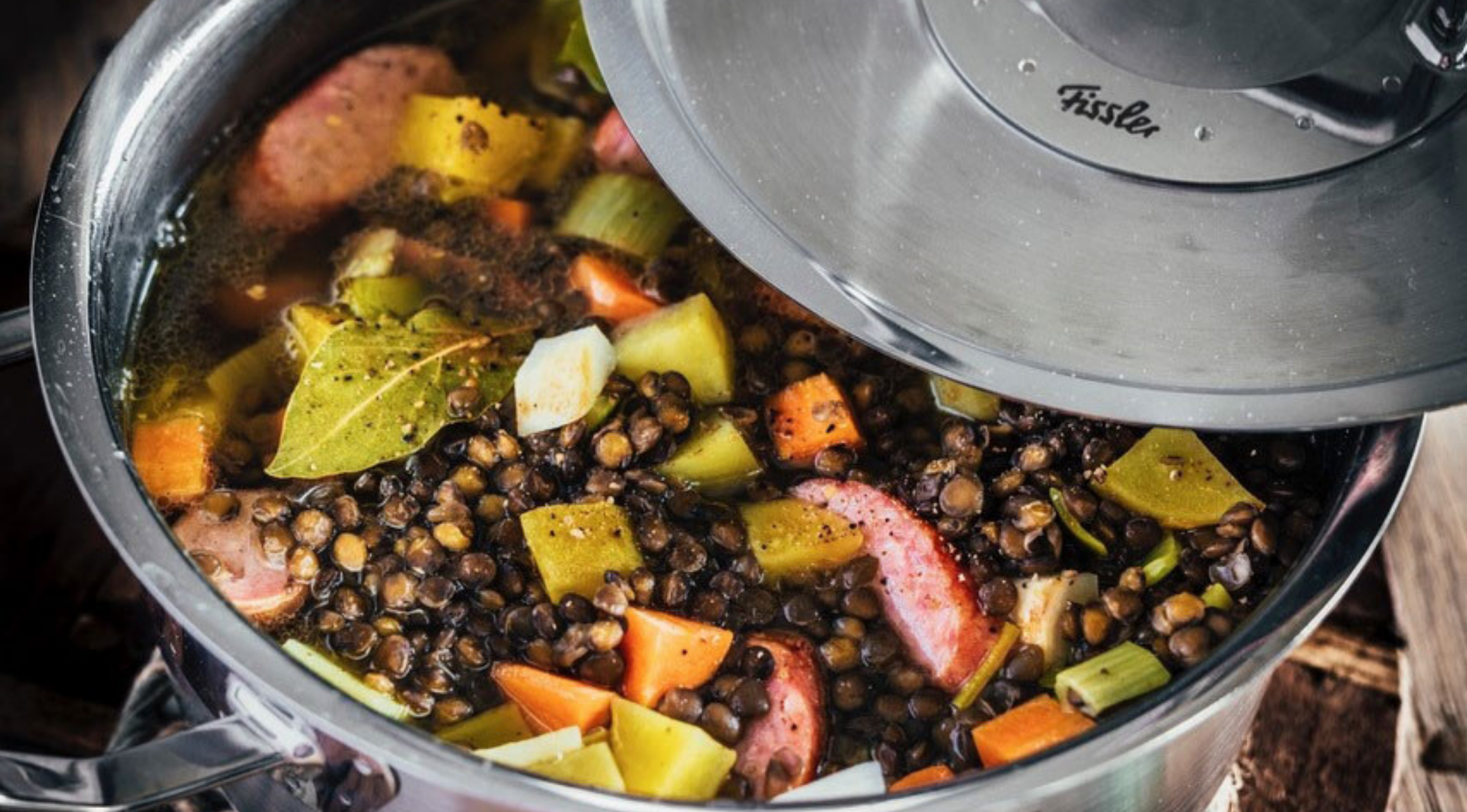 In einem Fissler-Edelstahltopf mit angehobenem Deckel wird Linsensuppe mit Wurst, Karotten, Kartoffeln, Sellerie und einem Lorbeerblatt serviert.
