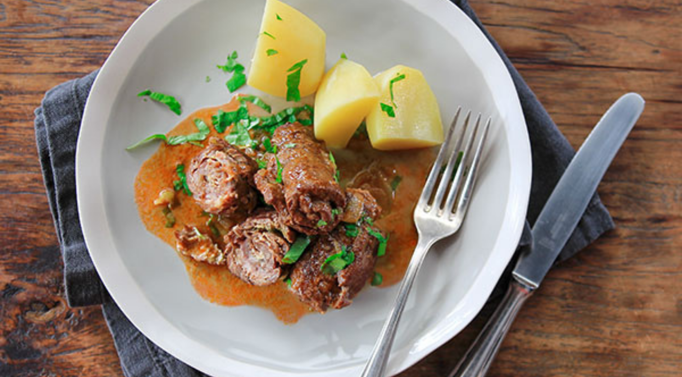Rinderrouladen in reicher brauner Sauce mit Kartoffeln auf einem weißen Teller, serviert mit Fissler-Besteck auf einem Holztisch.