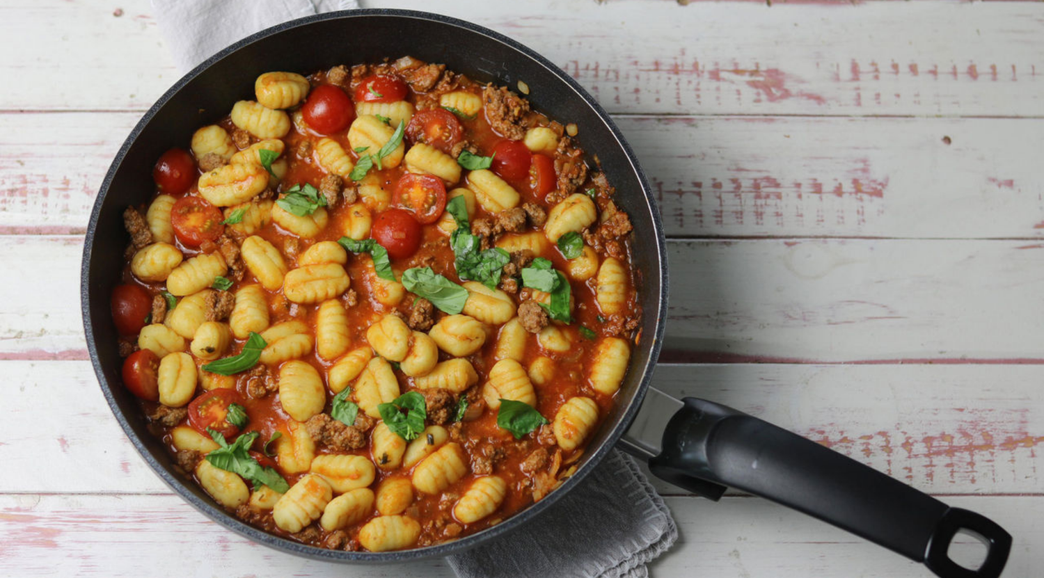 Eine hochwertige Fissler-Pfanne mit Gnocchi, Fleisch, Kirschtomaten und Basilikum in Tomatensauce auf Holzoberfläche mit grauem Tuch.