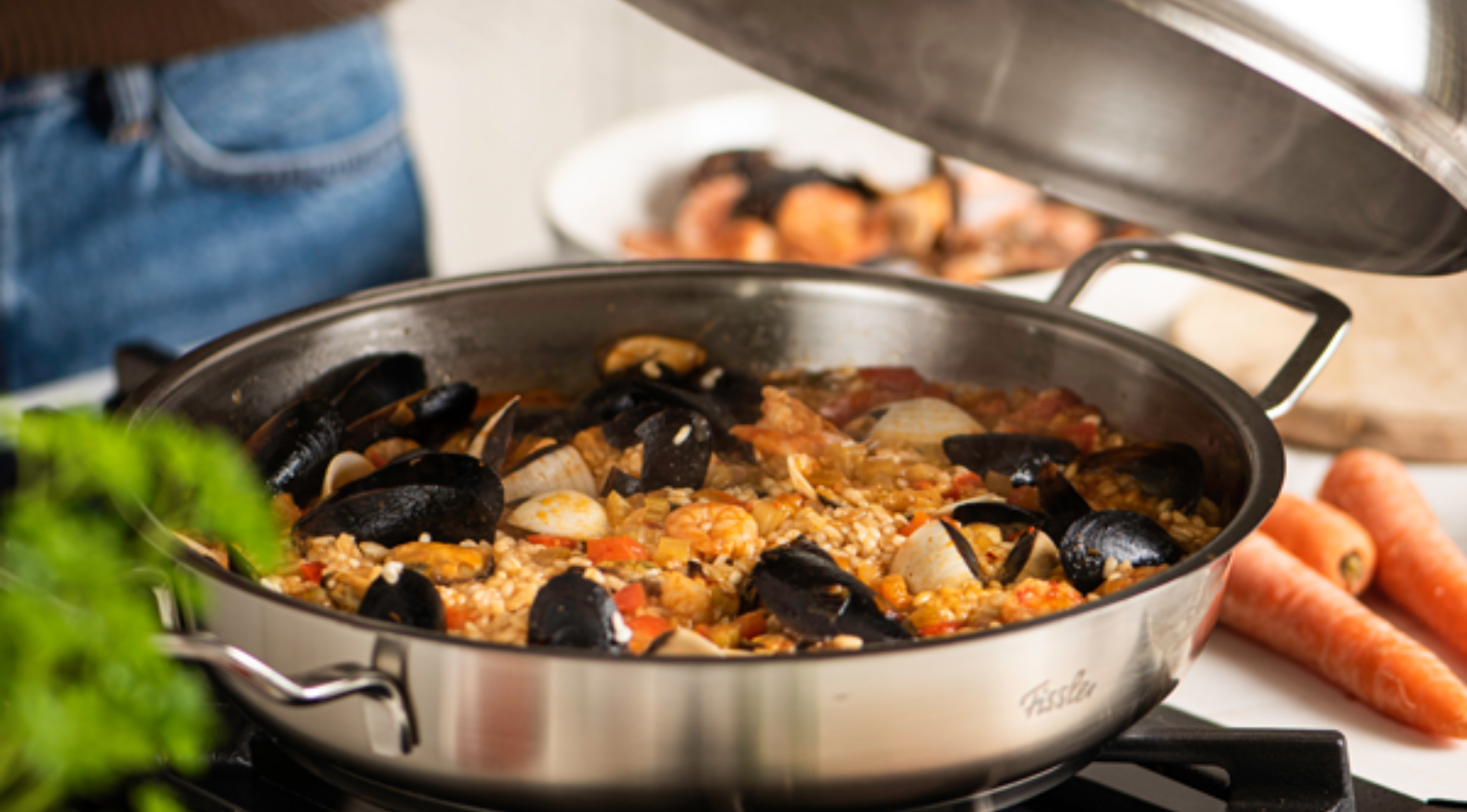 Eine Fissler-Edelstahlpfanne mit Paella mit Meeresfrüchten, eine Hand hebt den passgenauen Deckel an; frische Karotten und Grünzeug auf der Theke.