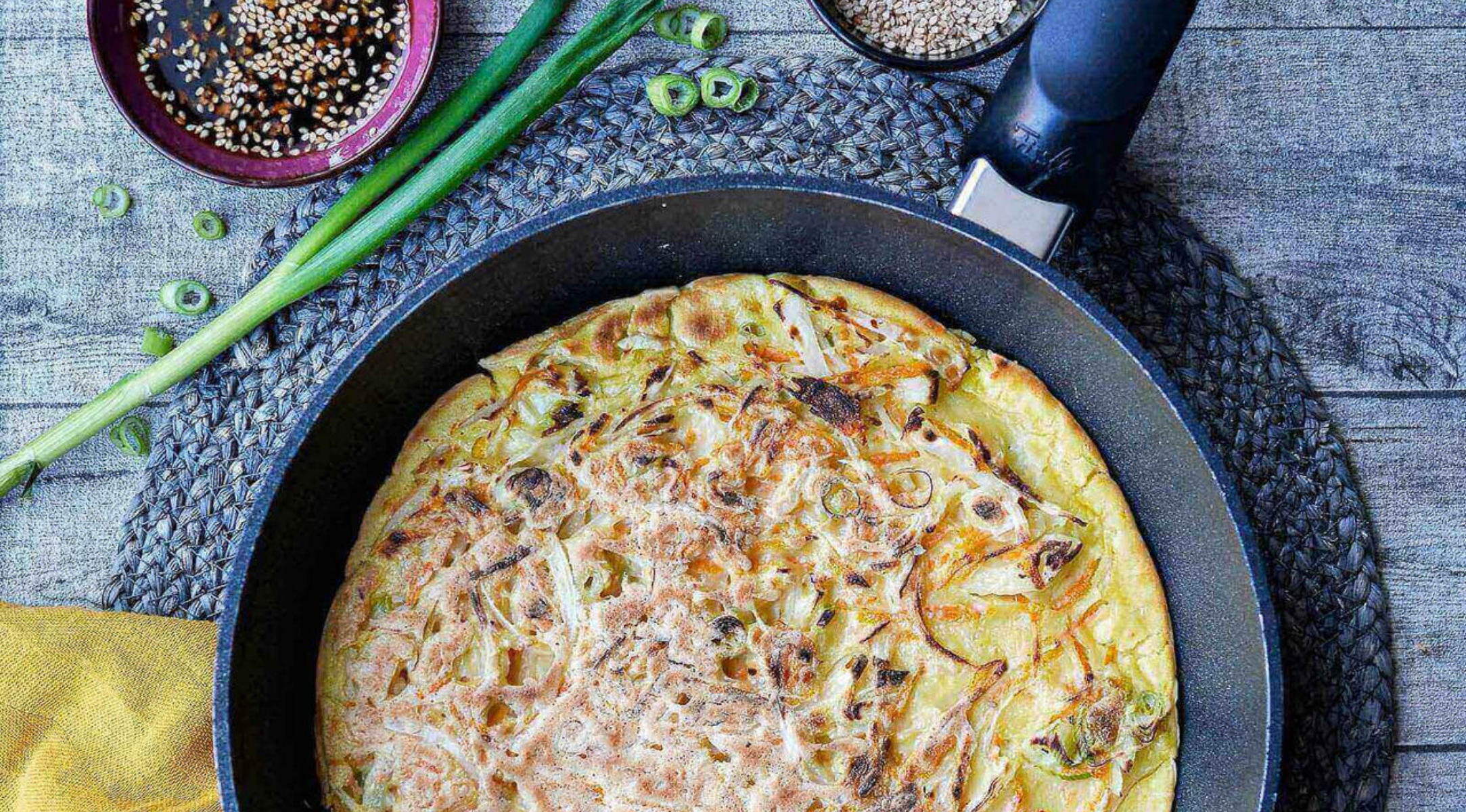 Ein koreanisches Pajeon mit grünen Zwiebeln wird in einer Fissler-Pfanne gegart, die mit frischen Belägen und Soßen auf einem Holztisch steht.