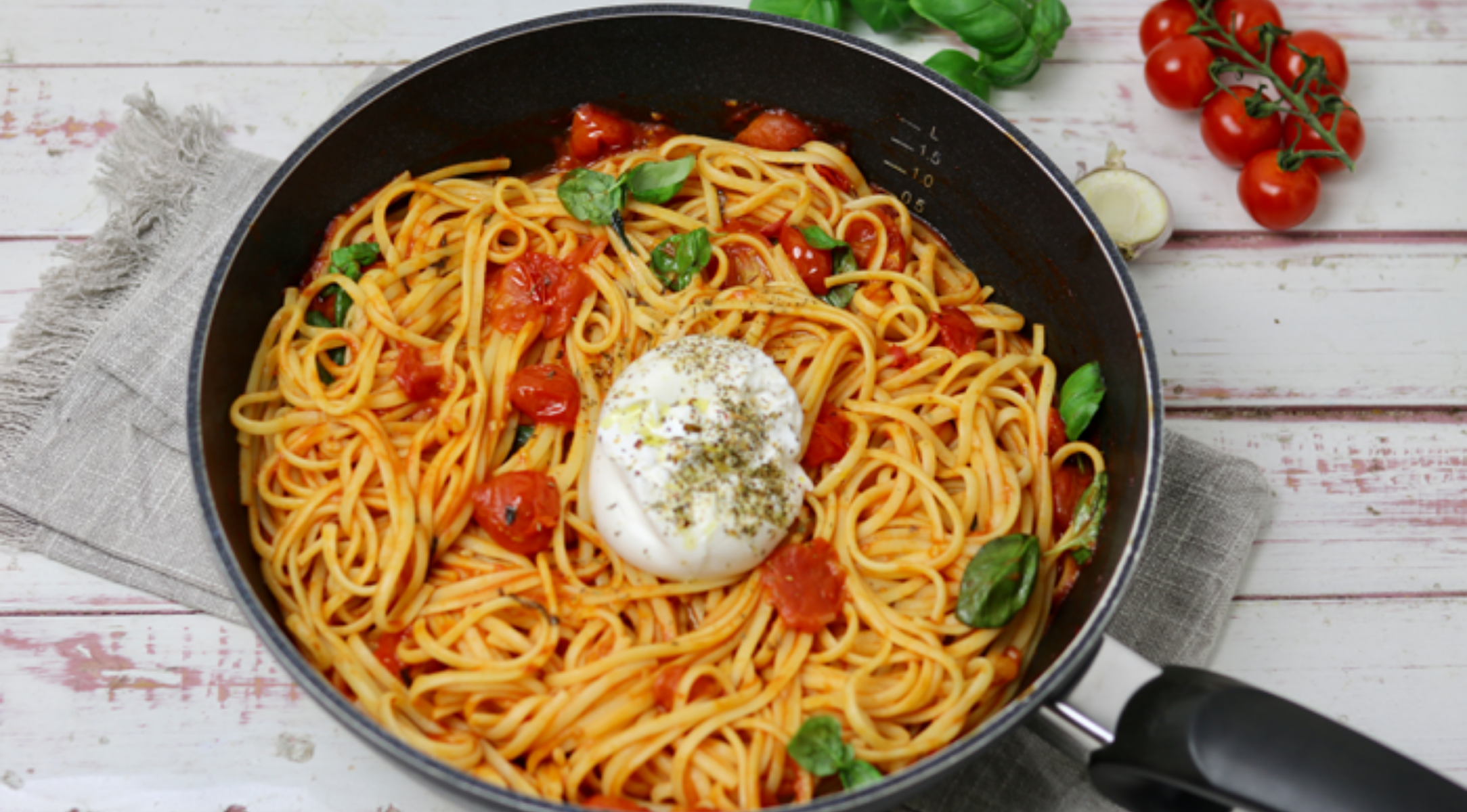 Spaghetti in Tomatensauce mit Basilikum und Burrata, serviert in einer hochwertigen Fissler-Pfanne auf einem rustikalen Tisch, daneben Tomaten und Kräuter.