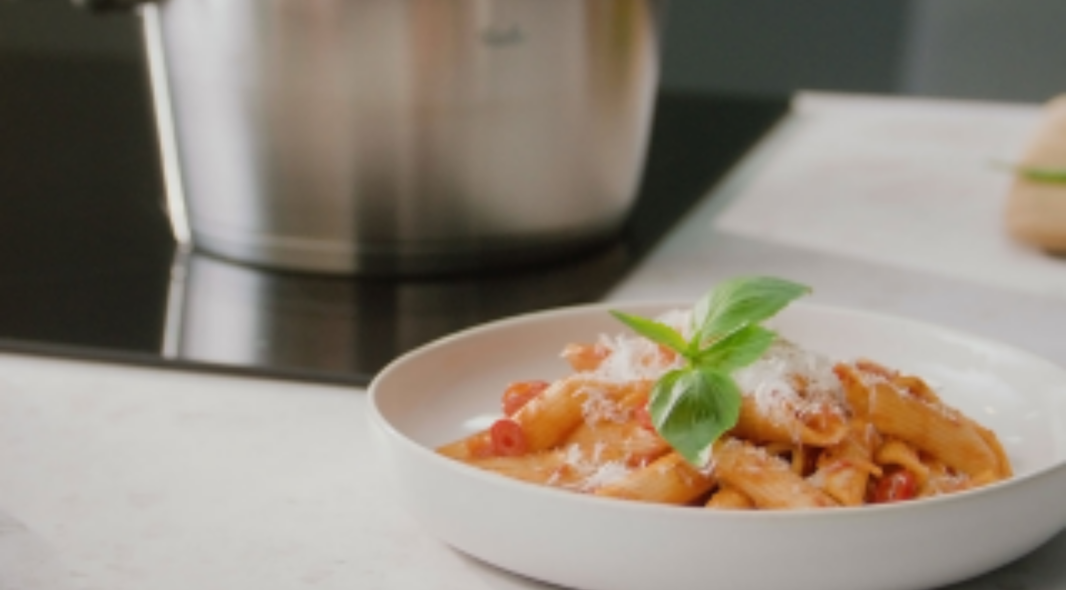 Penne mit Tomatensauce, Käse und Basilikum auf einem Teller neben einem Fissler-Edelstahltopf auf der Arbeitsplatte.