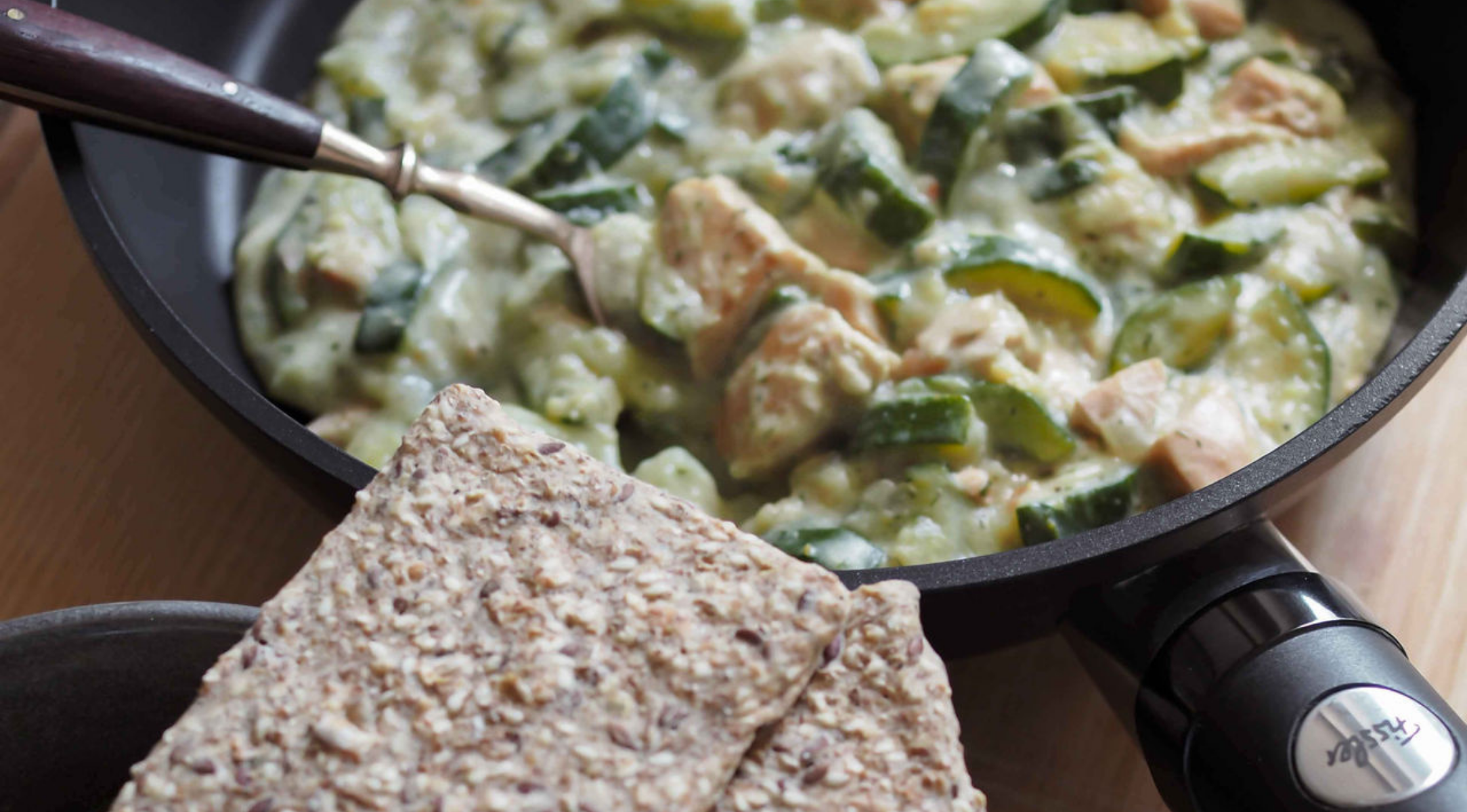 Eine Fissler-Pfanne mit cremigem Hähnchen und Zucchini neben Knäckebrot auf einem Teller; eine Gabel ruht in der hochwertigen Edelstahlpfanne.