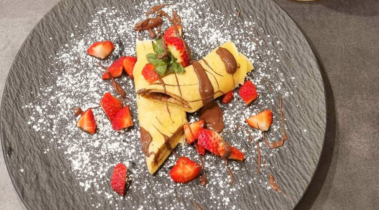 Zwei gerollte Crêpes mit Schokostreuseln, Erdbeeren und Minze auf einem dunklen Teller, elegant präsentiert auf hochwertigem Fissler-Geschirr.