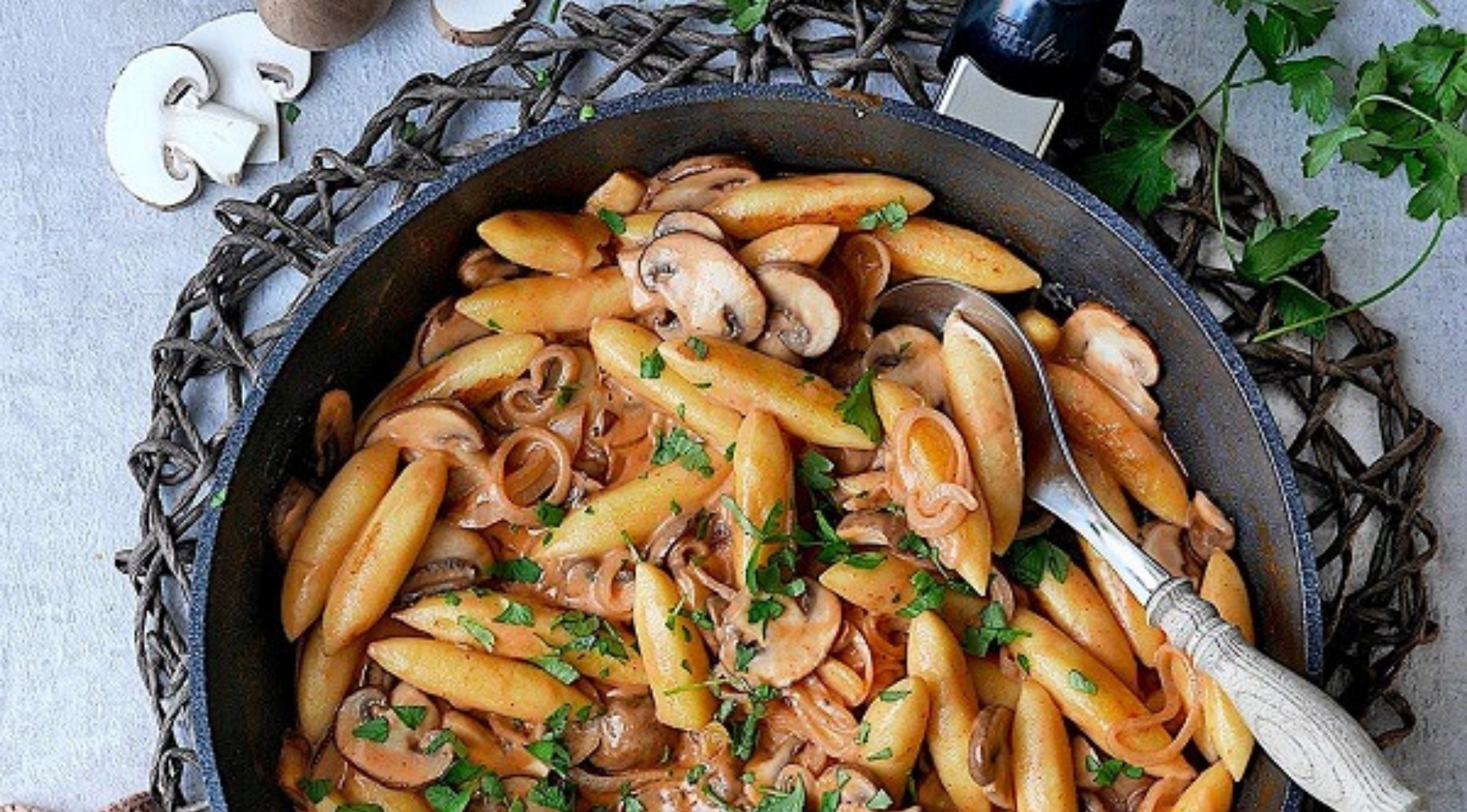 Eine Fissler-Edelstahlpfanne mit Nudeln, Champignons, Zwiebeln, Petersilie; darin ein Löffel. Rohe Champignons und Kräuter neben der Pfanne.