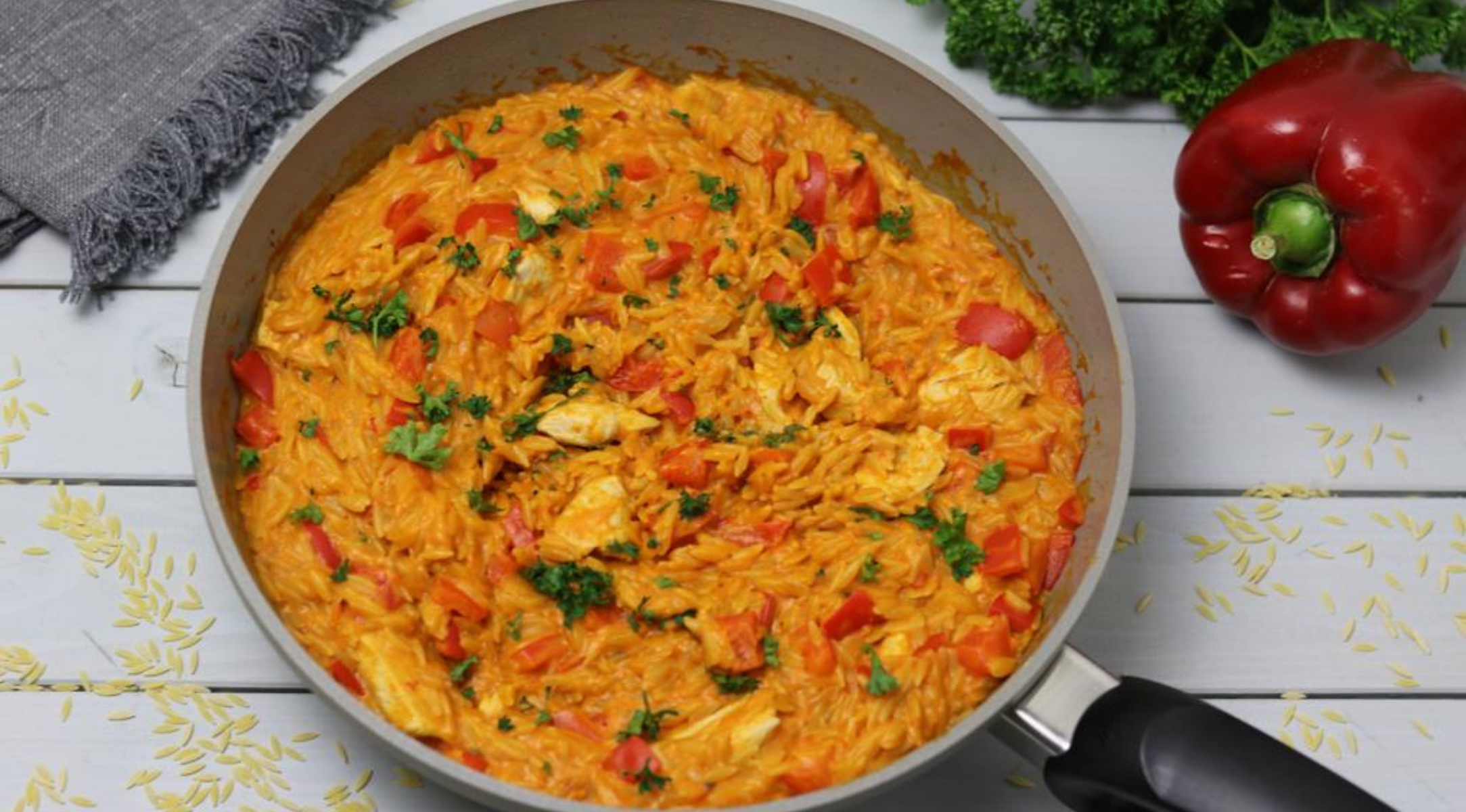 Fissler-Pfanne mit cremigem Orzo, Huhn, rotem Paprika und Kräutern. Graue Serviette und Gemüse auf einem weißen Holztisch.