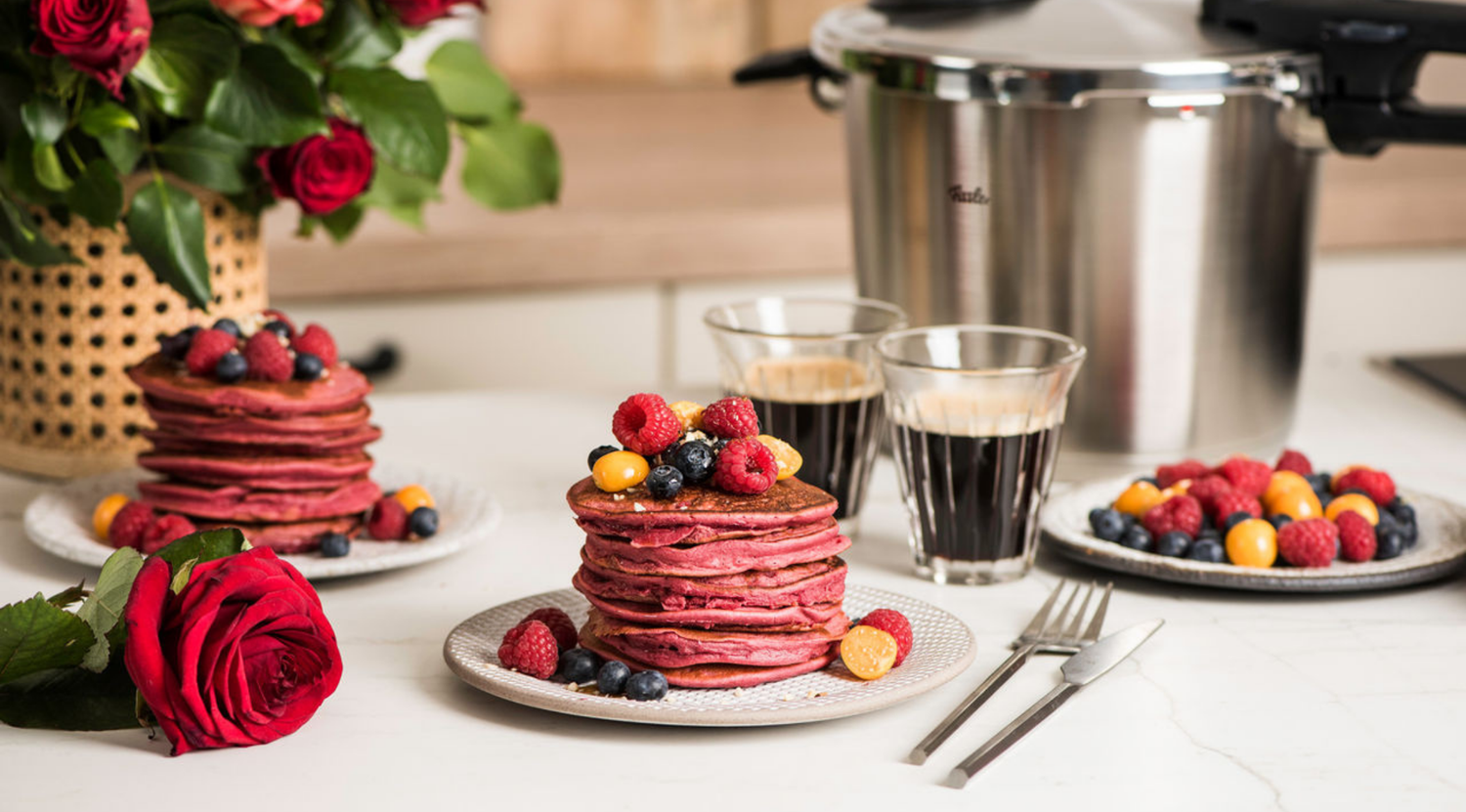 Elegante Küchenszene: Fissler Schnellkochtopf, rote Pfannkuchen mit Beeren, Kaffee, Rosen zeigen Qualität und zeitloses Design.