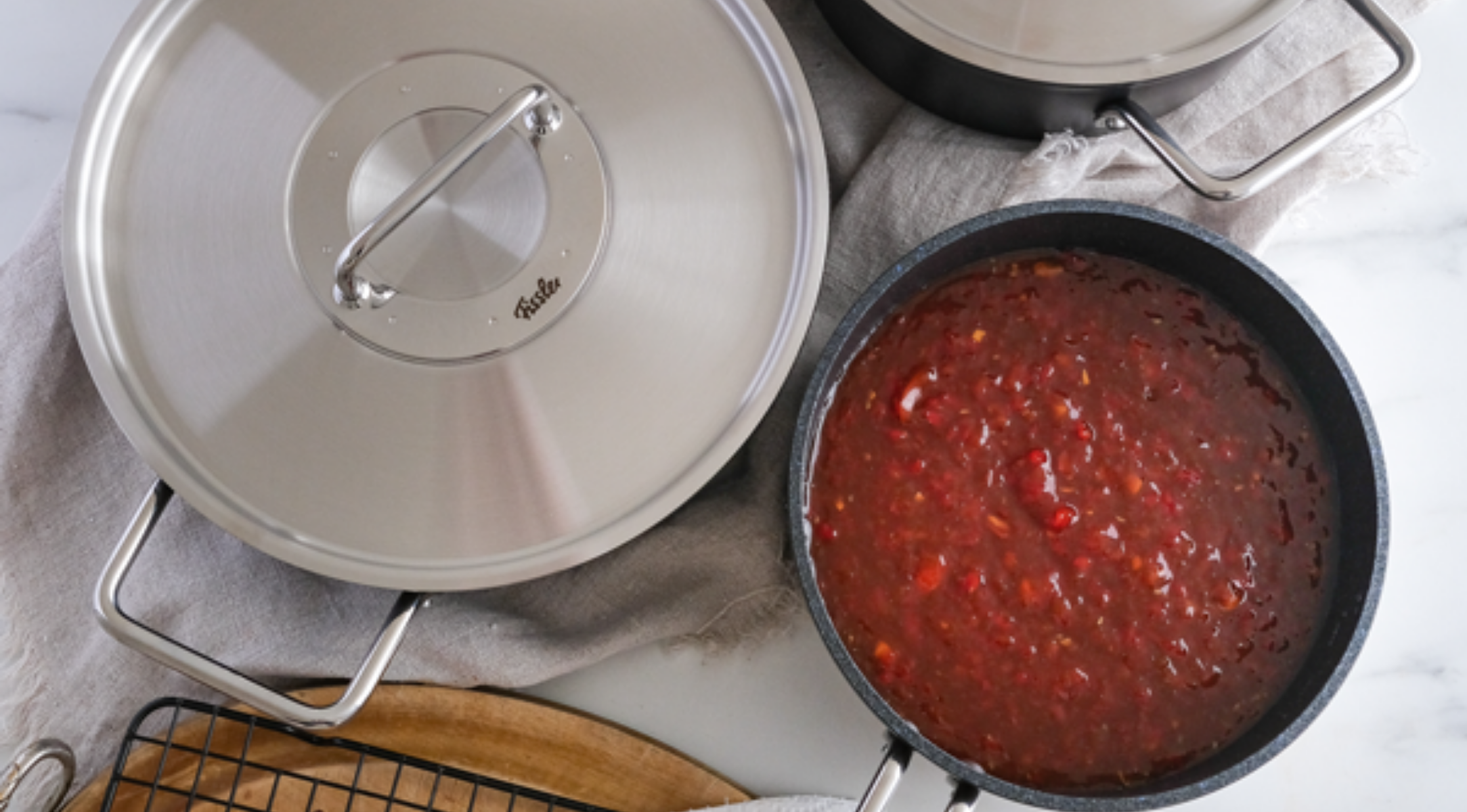 Töpfe aus rostfreiem Stahl von Fissler, Präzisionsarbeit aus Deutschland, neben einem Topf mit stückiger Tomatensauce auf einem hellen Tuch auf einer Marmorarbeitsplatte.