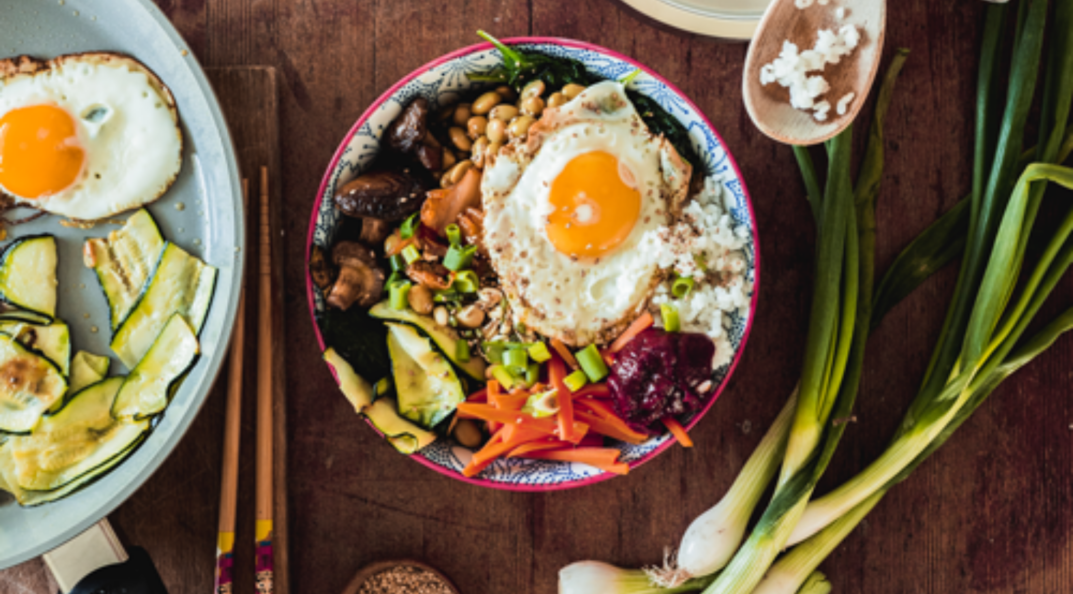 Eine lebhafte Schüssel Bibimbap neben frischem Gemüse und einer Fissler-Pfanne mit Spiegelei, alles auf einem Holztisch mit Stäbchen.