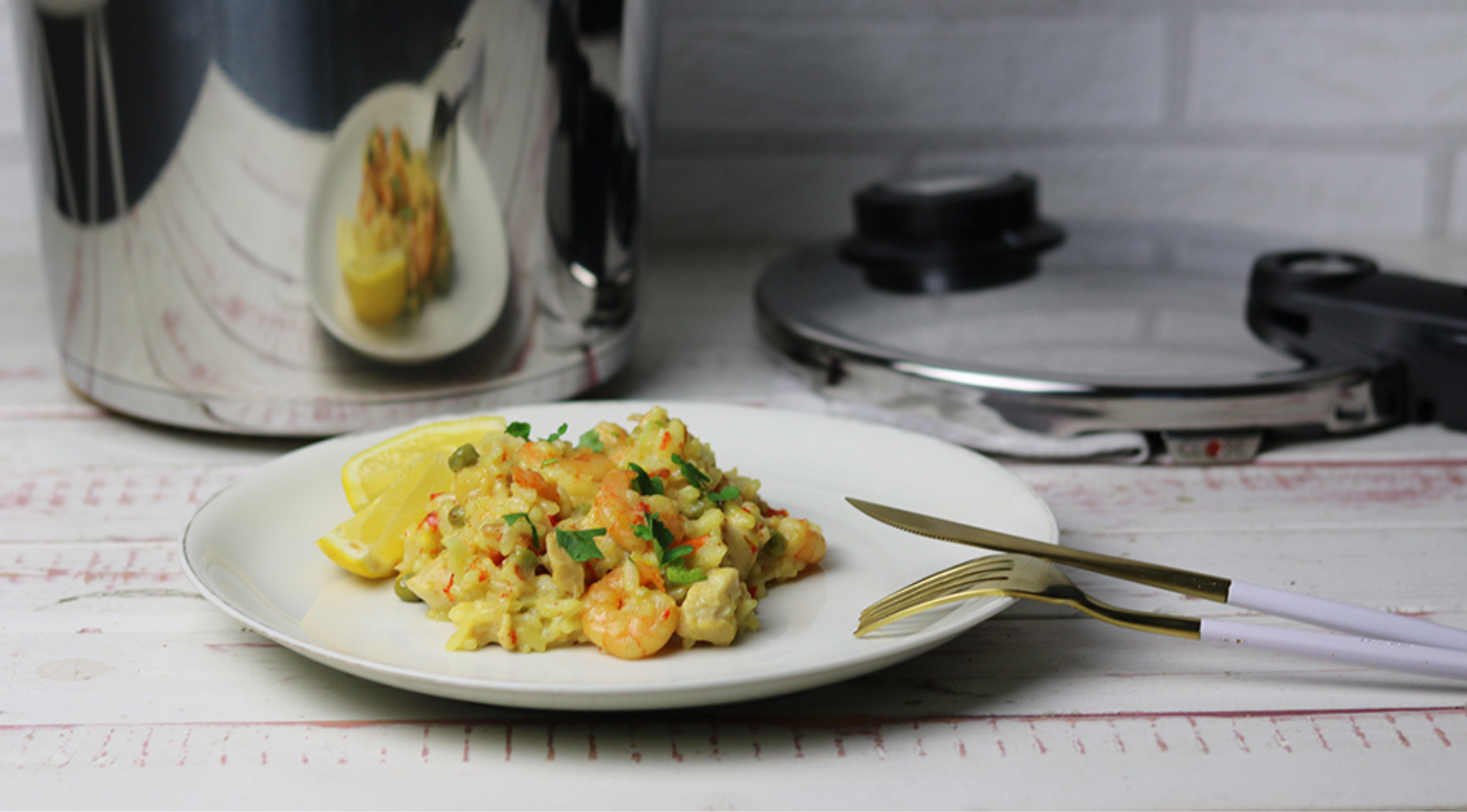 Cremige Garnelen und Reis, Kräuter und Zitrone auf einem Tisch mit goldenem Besteck; im Hintergrund ein Fissler Schnellkochtopf aus Edelstahl.