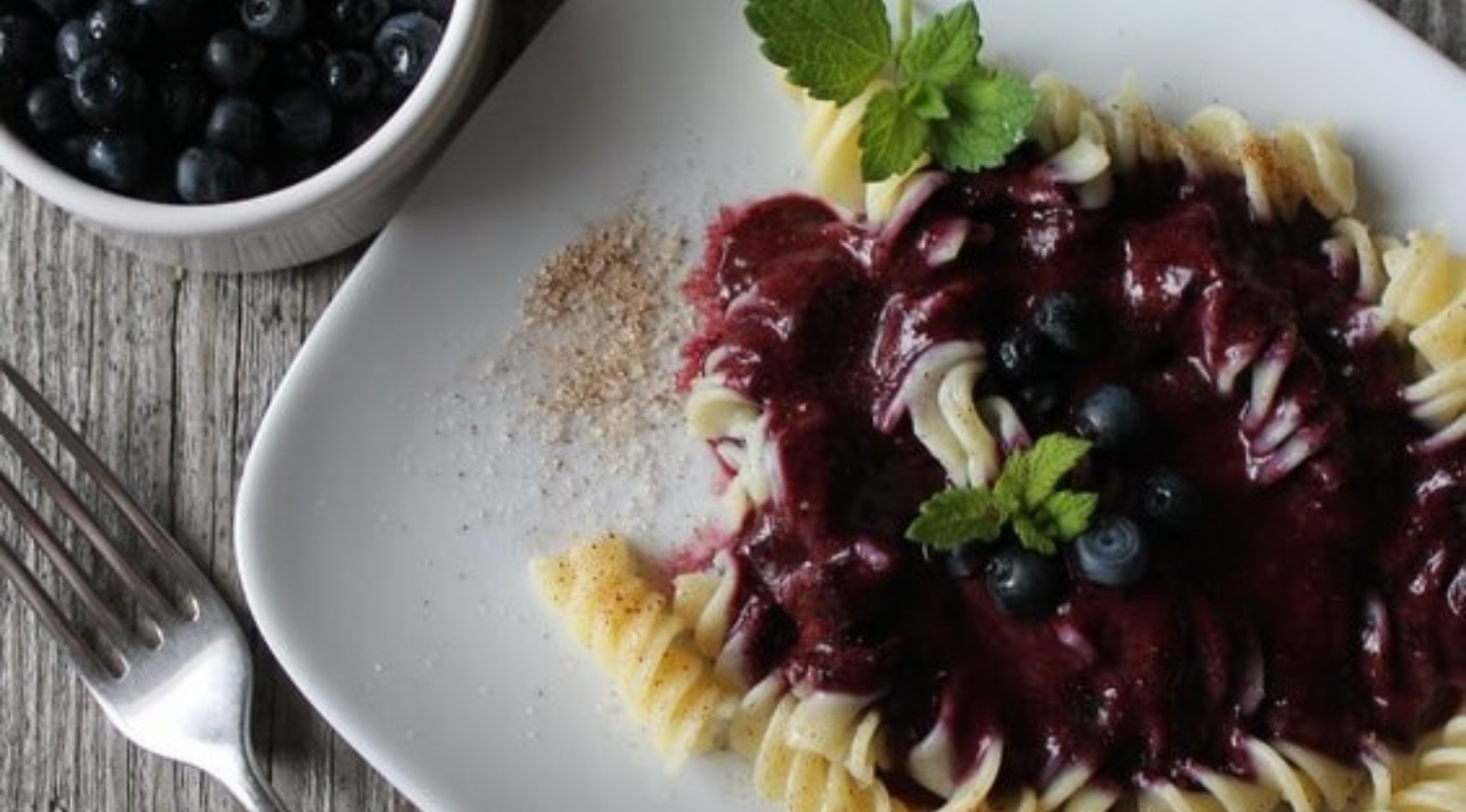 Rotini-Nudeln mit Blaubeersauce und Minze auf einem Holztisch, serviert mit einer Fissler-Gabel und einer Schale frischer Blaubeeren.