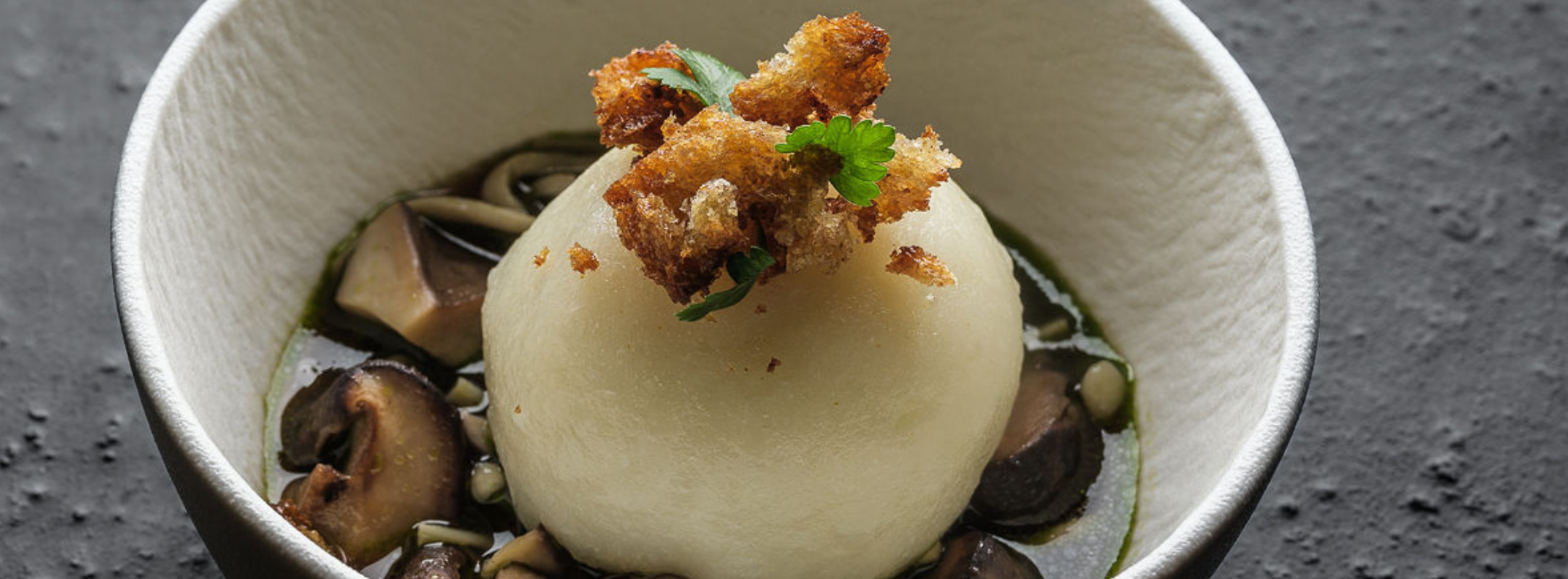 A perfectly round dumpling rests in a white bowl filled with clear broth, encircled by delicately sliced mushrooms and crowned with crispy garnishes and a fresh parsley sprig—prepared and served using Fissler’s precision-engineered cookware for outstanding results.