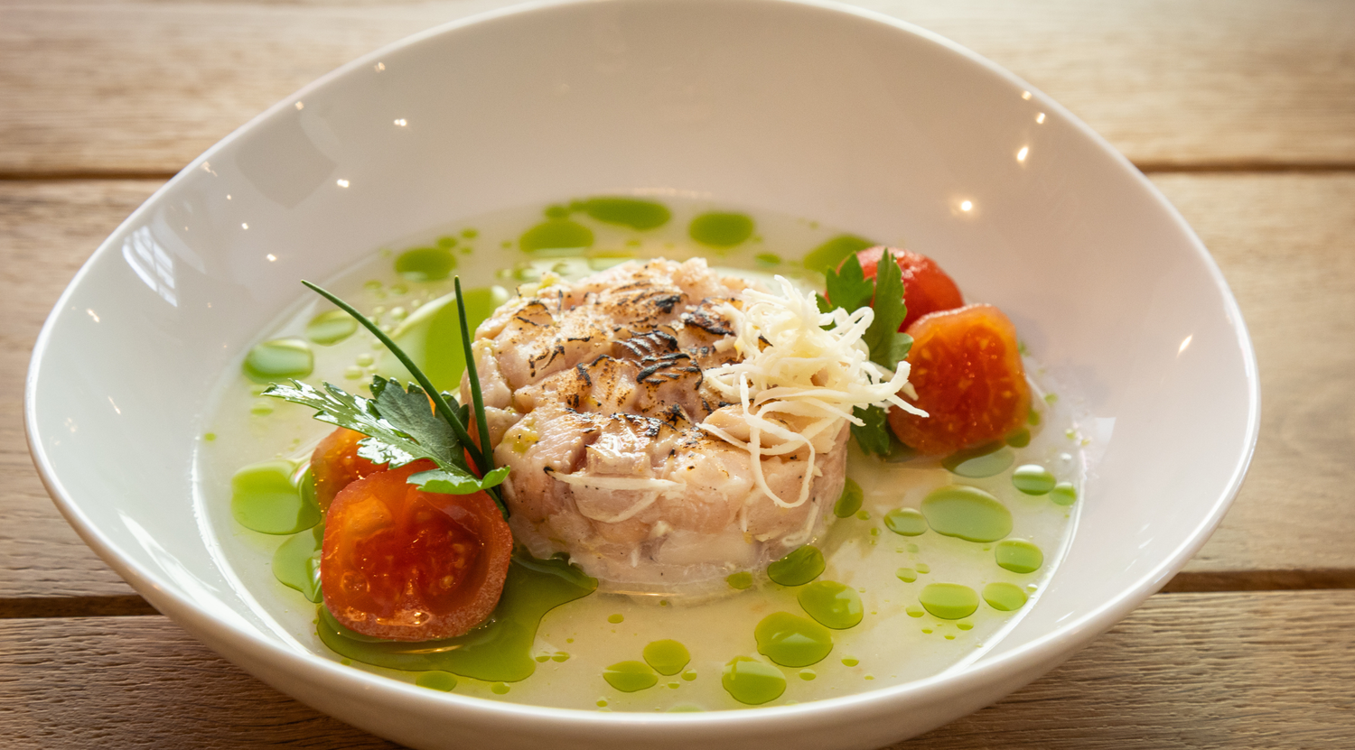 Eine Gourmet-Fischkreation, elegant angerichtet in einer flachen weißen Schale, garniert mit geriebenem Käse, halbierten Kirschtomaten, Petersilie und leuchtend grünen Öltröpfchen. Das raffinierte Gericht steht auf einem Holztisch und verdeutlicht die Präzision, die mit dem hochwertigen Edelstahl-Kochgeschirr von Fissler erreicht werden kann – wobei deutsche Ingenieurskunst außergewöhnliche Qualität für jedes kulinarische Meisterwerk gewährleistet.