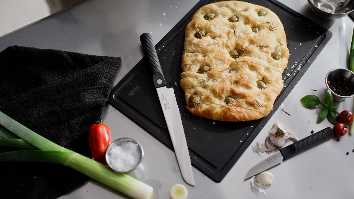 A rectangular focaccia on a board next to the Fissler ProCut bread knife 21cm, fresh produce and kitchen accents on a gray surface.