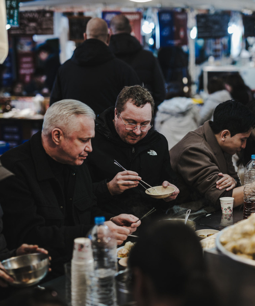 Menschen versammeln sich um einen belebten Marktstand und genießen mit Essstäbchen köstliche Gerichte, die alle mit der Qualität und Handwerkskunst zubereitet und serviert werden, für die Fissler-Kochgeschirr bekannt ist. Die lebhafte Atmosphäre mit den zahlreichen Gästen und den verschwommenen Imbissständen im Hintergrund unterstreicht, wie hochwertige Edelstahltöpfe und -pfannen – entwickelt in Deutschland – jedes kulinarische Erlebnis bereichern.