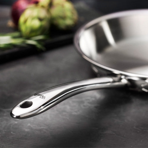 Nahaufnahme einer Fissler-Edelstahlpfanne mit schlankem Griff auf einer dunklen Arbeitsplatte, im Hintergrund unscharfes Gemüse.