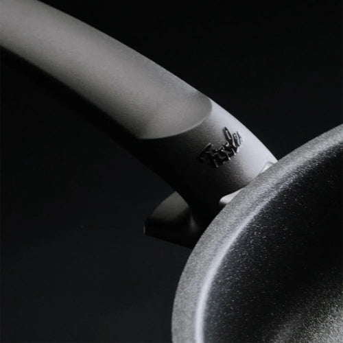 Nahaufnahme einer Fissler Bratpfanne mit gebogenem Griff und eingeprägtem Markenzeichen. Der dunkle Hintergrund unterstreicht das Design und die Qualität deutscher Ingenieurskunst.