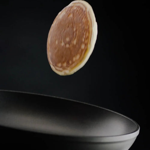 Ein goldener Pfannkuchen schwenkt über einer Fissler Premium-Bratpfanne, die deutsche Präzision und zeitloses Design vor einem dunklen Hintergrund hervorhebt.