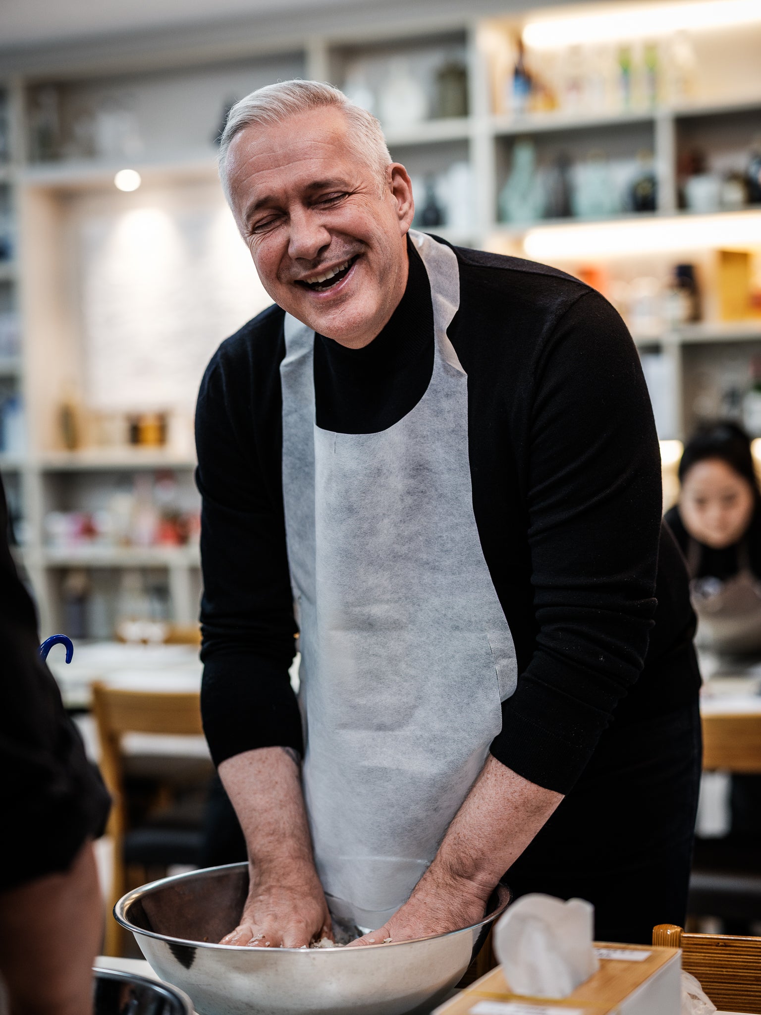 A smiling man in a black shirt and white apron kneads dough in a large Fissler bowl at a cooking class, surrounded by shelves and other people. The scene highlights the use of premium, German-engineered Fissler cookware, reflecting precision and timeless design in a modern kitchen setting.