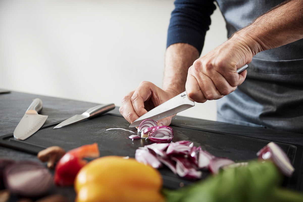 Eine Person schneidet eine rote Zwiebel präzise mit dem Fissler Essential® Universalmesser, neben dem charakteristischen frischen Gemüse und dem klaren Design.