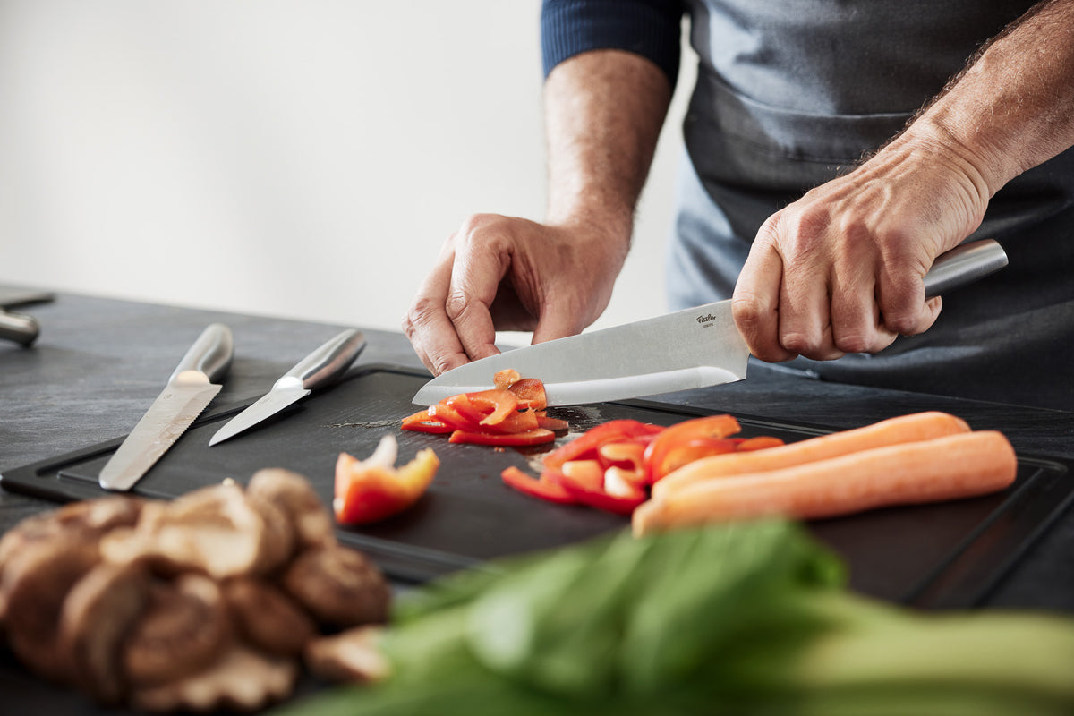 Ein Koch schneidet eine rote Paprika mit dem Fissler Essential® Kochmesser, umgeben von frischem Gemüse und eleganten Fissler-Messern.