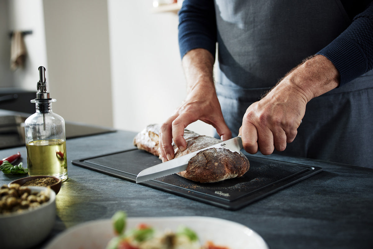 Eine Person in einer Schürze schneidet Brot mit dem Fissler Essential® Brotmesser auf einem Brett; Fissler Kochgeschirr setzt Akzente in der modernen Küche.