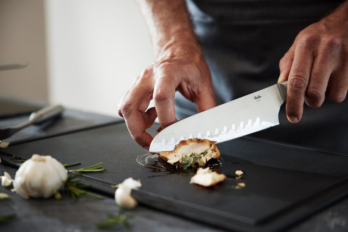 Schneiden von gefülltem Huhn mit dem Fissler Essential® Santokumesser auf einem schwarzen Brett, flankiert von Knoblauch und Rosmarin.
