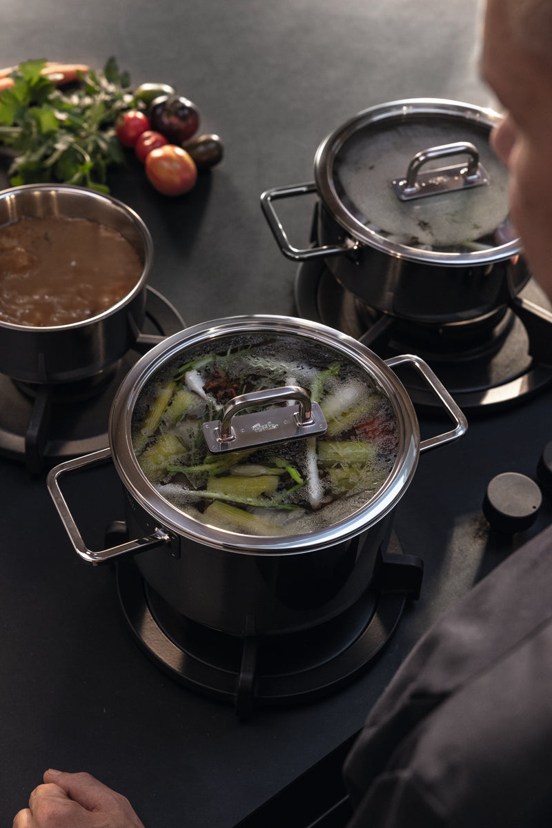 Eine Person kocht mit drei hochwertigen Fissler-Edelstahltöpfen; Brühe, köchelndes Gemüse und frische Produkte vervollständigen die Szene.
