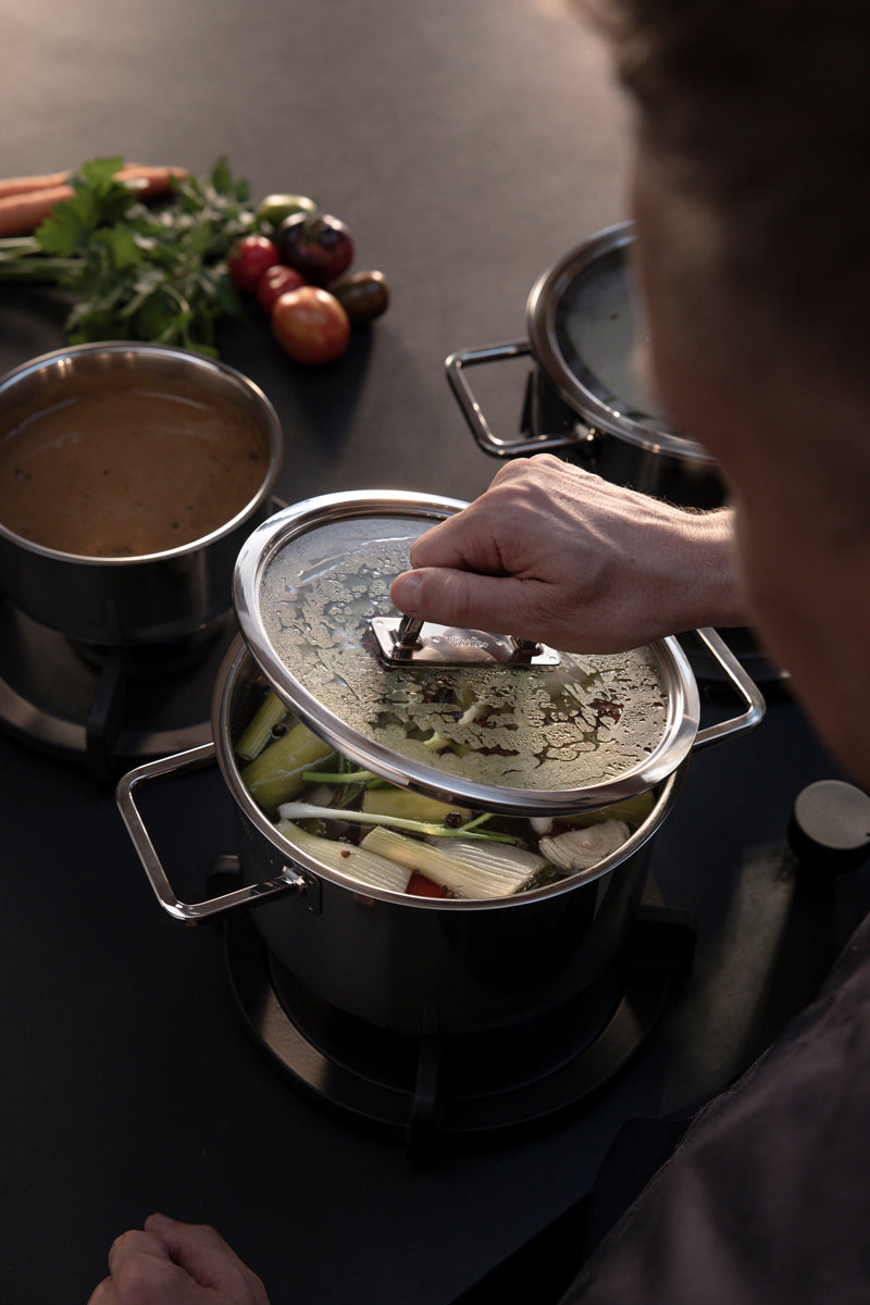 Eine Person hebt den Deckel eines mit Gemüse gefüllten Fissler Viseo® Pro-Edelstahltopfs an; die Soße köchelt und frisches Gemüse liegt daneben.