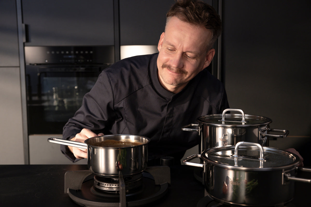 Ein Koch in einer modernen Küche genießt eine Sauce, die in einem Edelstahl Kochtopf köchelt, mit Fissler Viseo® Pro Töpfen in der Nähe.