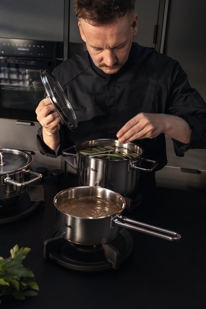 Ein Koch gibt grüne Bohnen in eine Fissler Viseo® Pro Stielkasserolle 16 cm; Edelstahltöpfe köcheln in einer eleganten, modernen Küche.