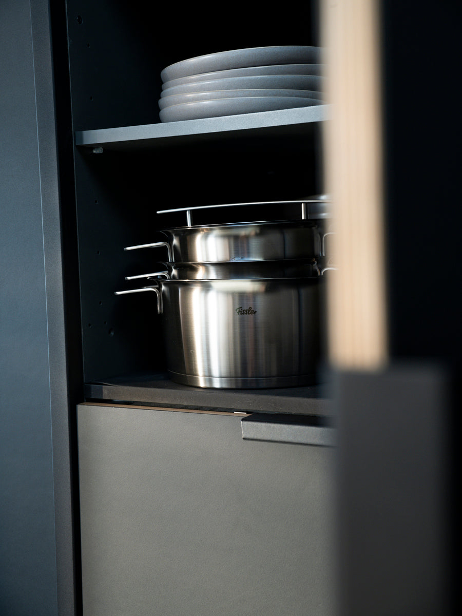 Elegant white plates are stacked above a Fissler Phi Collection® saucepan, showcasing timeless German-made design in a dark kitchen cabinet.