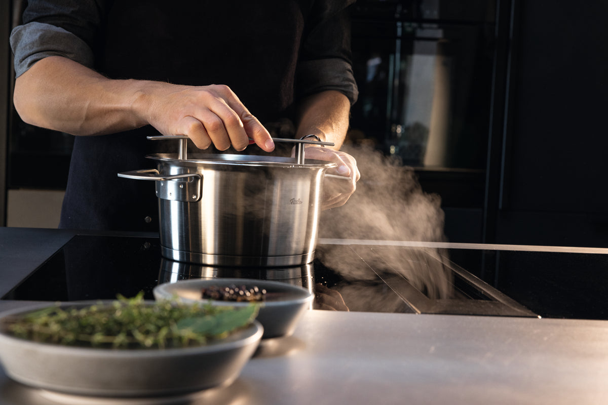Entdecken Sie die Präzision von Fissler: Beim Anheben des Deckels des Suppentopfs aus der Phi Collection® steigt Dampf auf; daneben liegen frische Kräuter und Pfefferkörner.