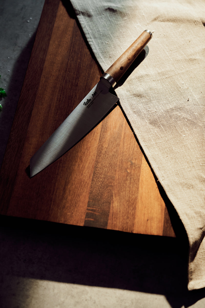 Fissler PrimeCut Kochmesser 20cm mit Holzgriff auf einem Brett, beiges Tuch, weiches Sonnenlicht-Präzision trifft auf zeitloses deutsches Design.