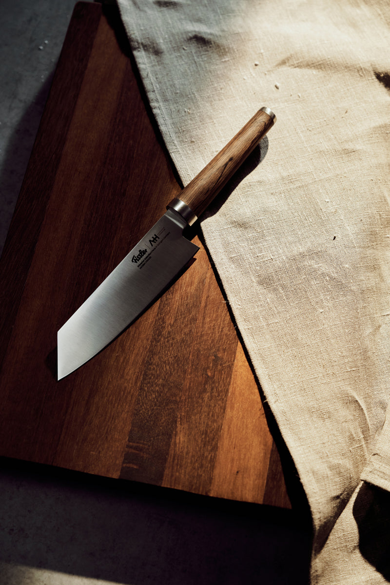 Ein Fissler-Kochmesser mit Holzgriff auf einem dunklen Brett, das teilweise von einem beigen Tuch umhüllt ist, beleuchtet von warmem Tageslicht.