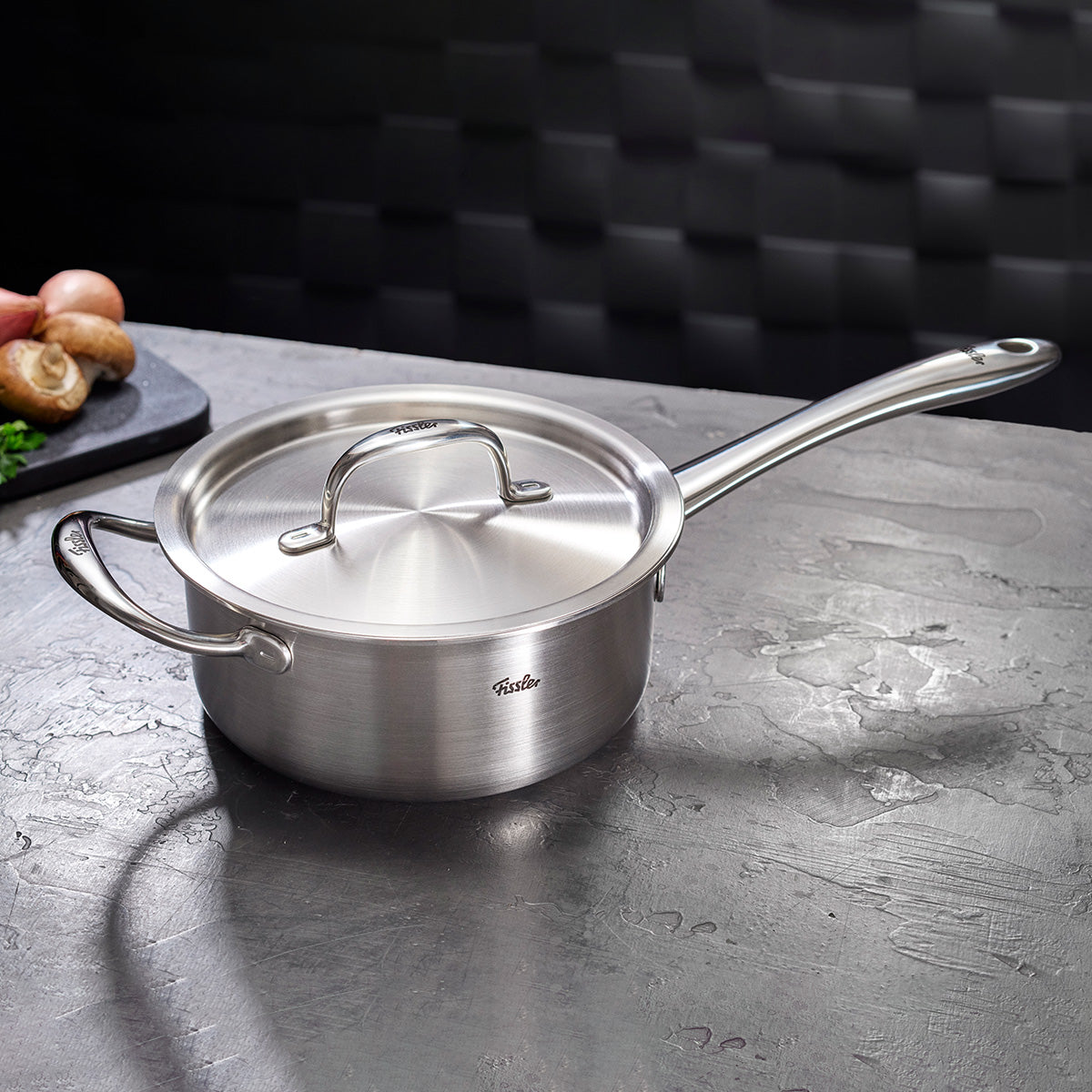 Die Fissler M5 Pro-Ply Stielkasserolle aus Edelstahl mit Deckel steht auf grauer Fläche, dahinter Brett mit Pilzen und Zwiebeln.