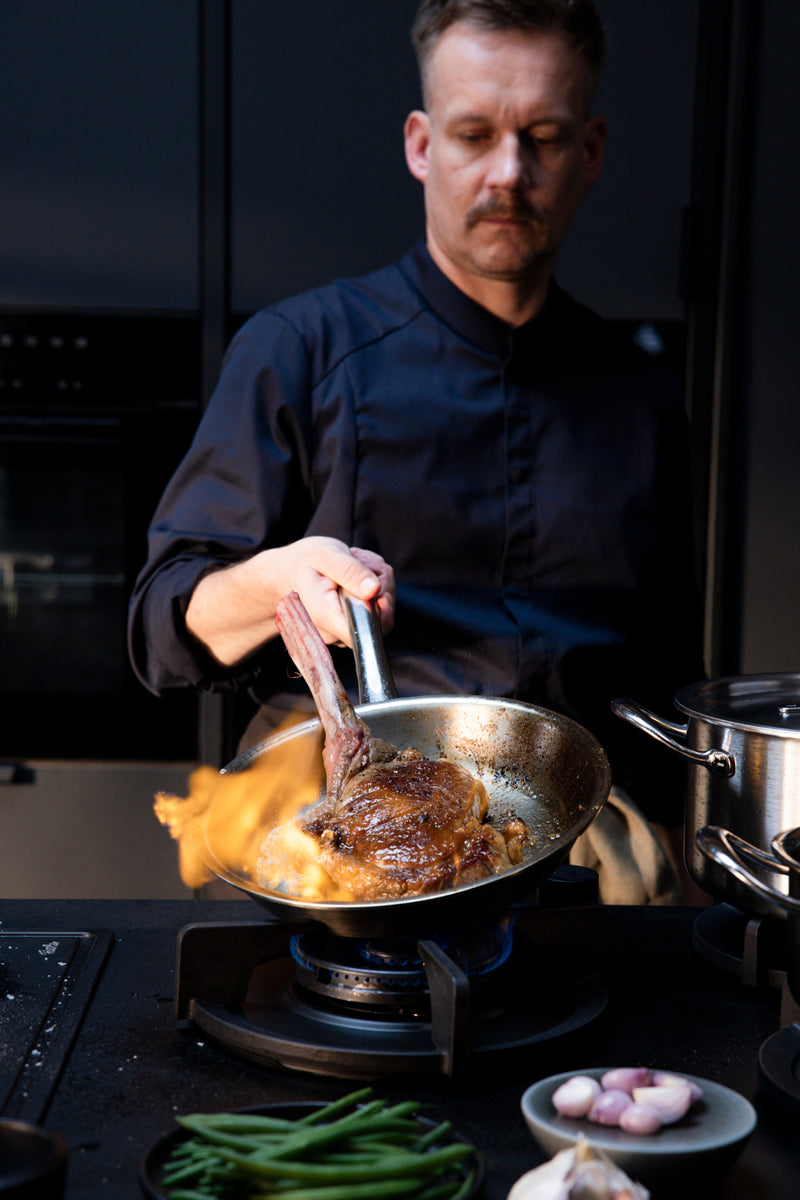 Ein Koch brät ein Steak fachmännisch in einer Fissler M5 Pro Ply® Pfanne an; Flammen steigen auf und zeigen die deutsche Qualität und Innovation.