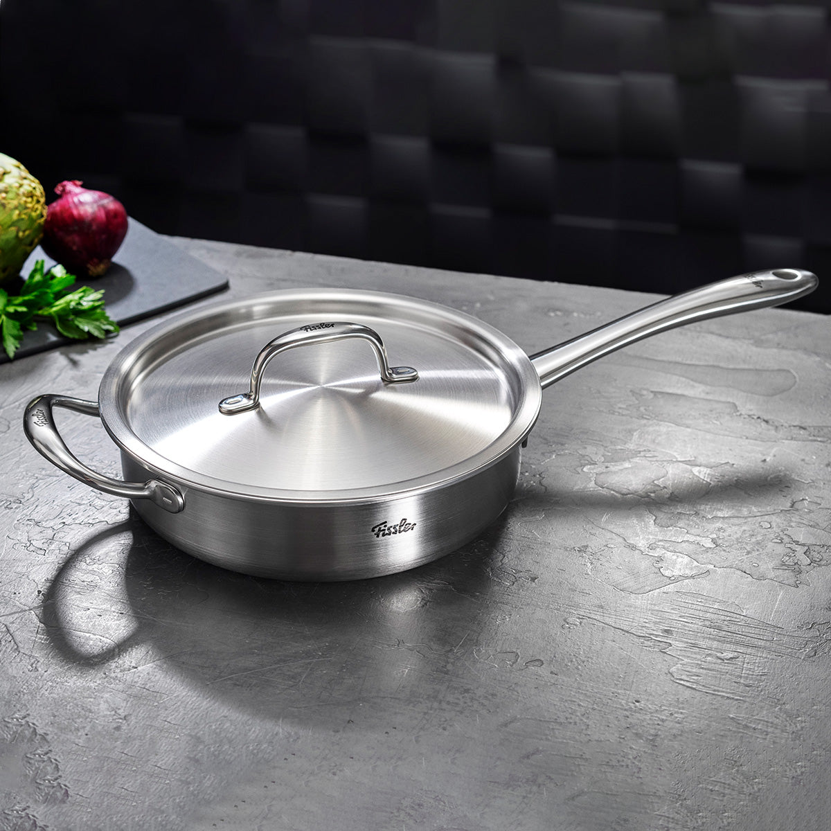 Fissler M5 Pro-Ply sauté pan made of recycled stainless steel on a gray plate, vegetables and artichoke in the background, suitable for induction.