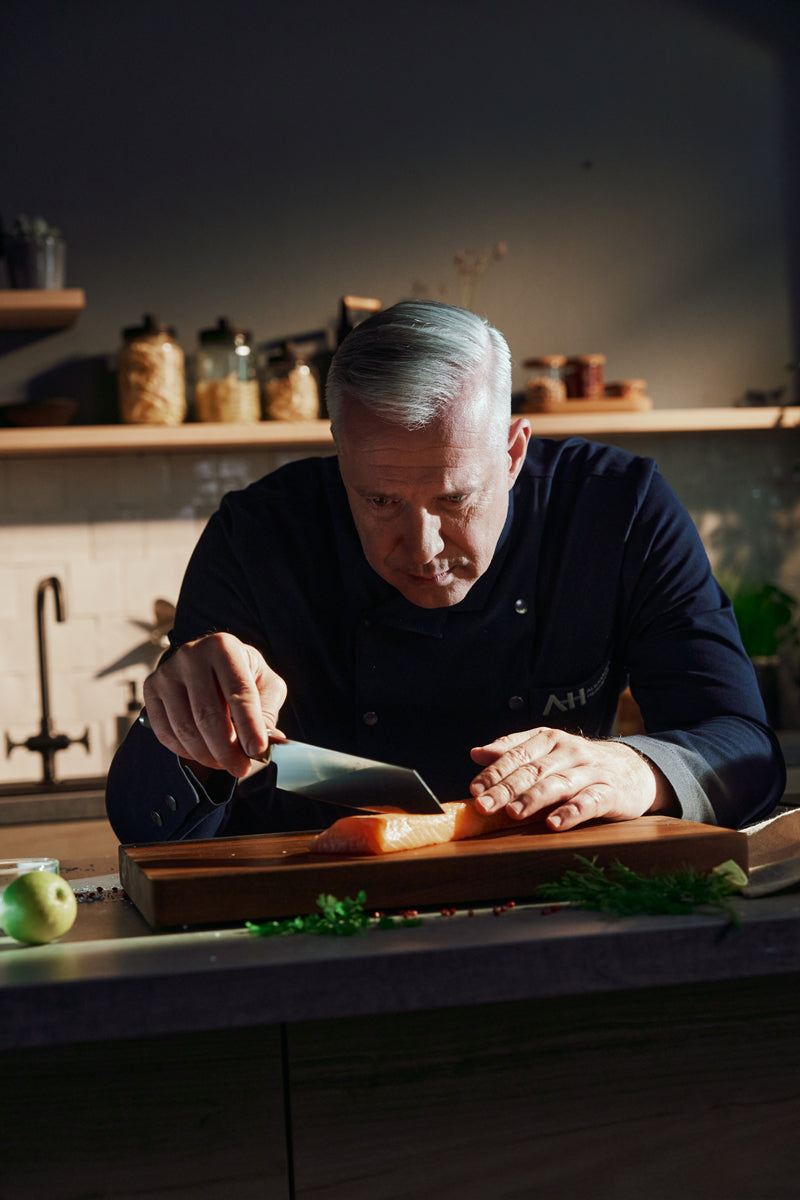 A chef uses the Fissler PrimeCut Santoku knife 17cm to slice salmon, with elegant glasses and utensils on modern kitchen shelves.