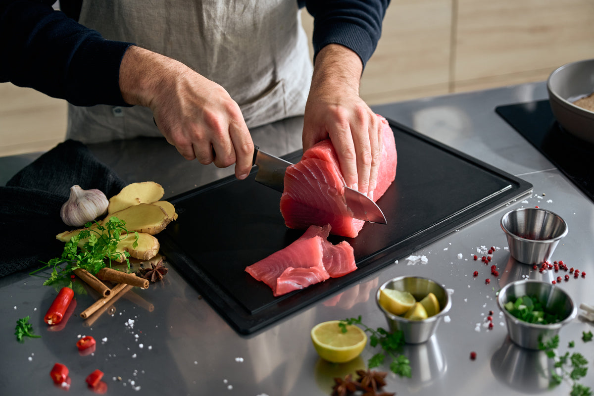 Die Zubereitung von frischem Thunfisch mit dem Fissler ProCut Santokumesser 17cm auf einem schlichten schwarzen Brett, inmitten von Kräutern und Gewürzen - Präzision made in Germany.