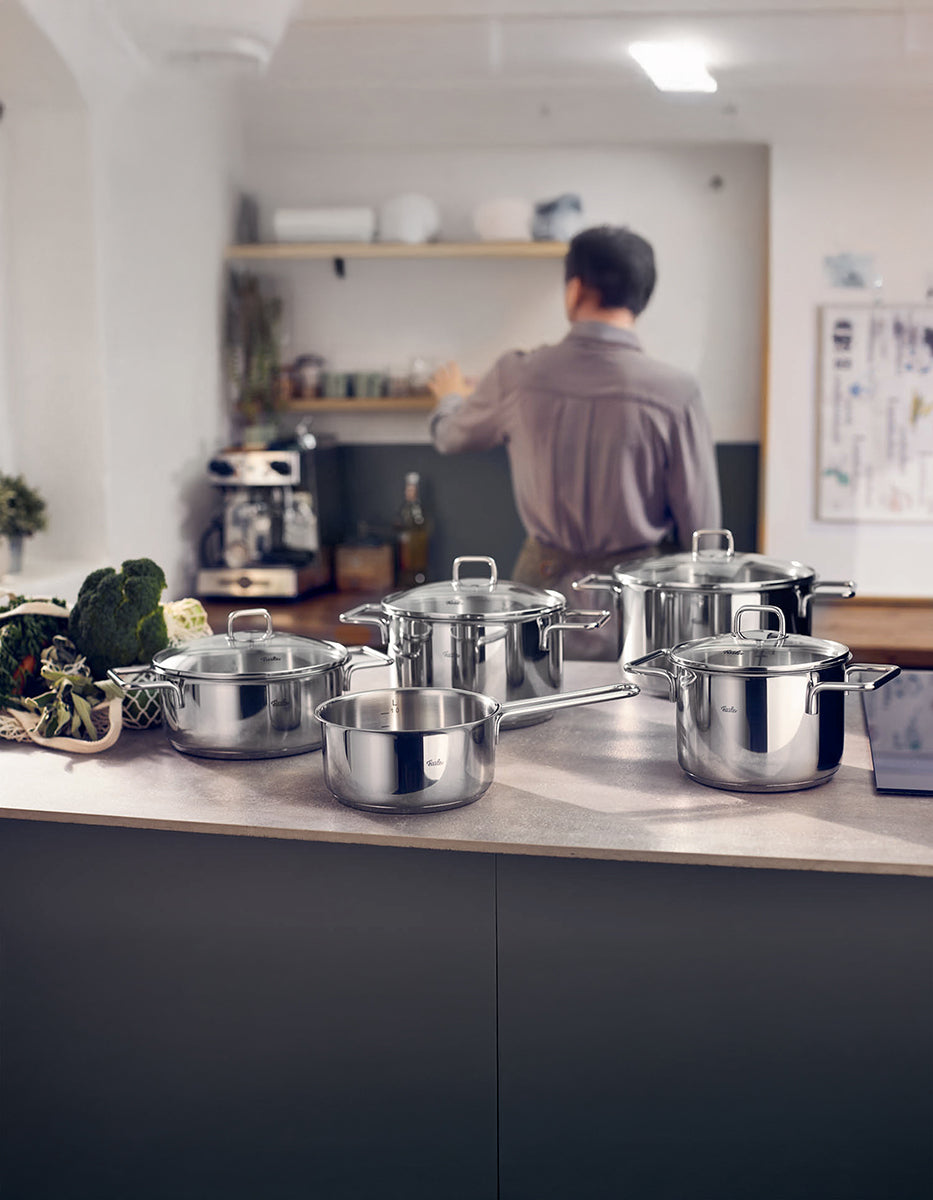 A kitchen with the 5-piece Fissler LumiTherm Cookware Set in stainless steel, fresh vegetables, and a person with shelves and a coffee machine.