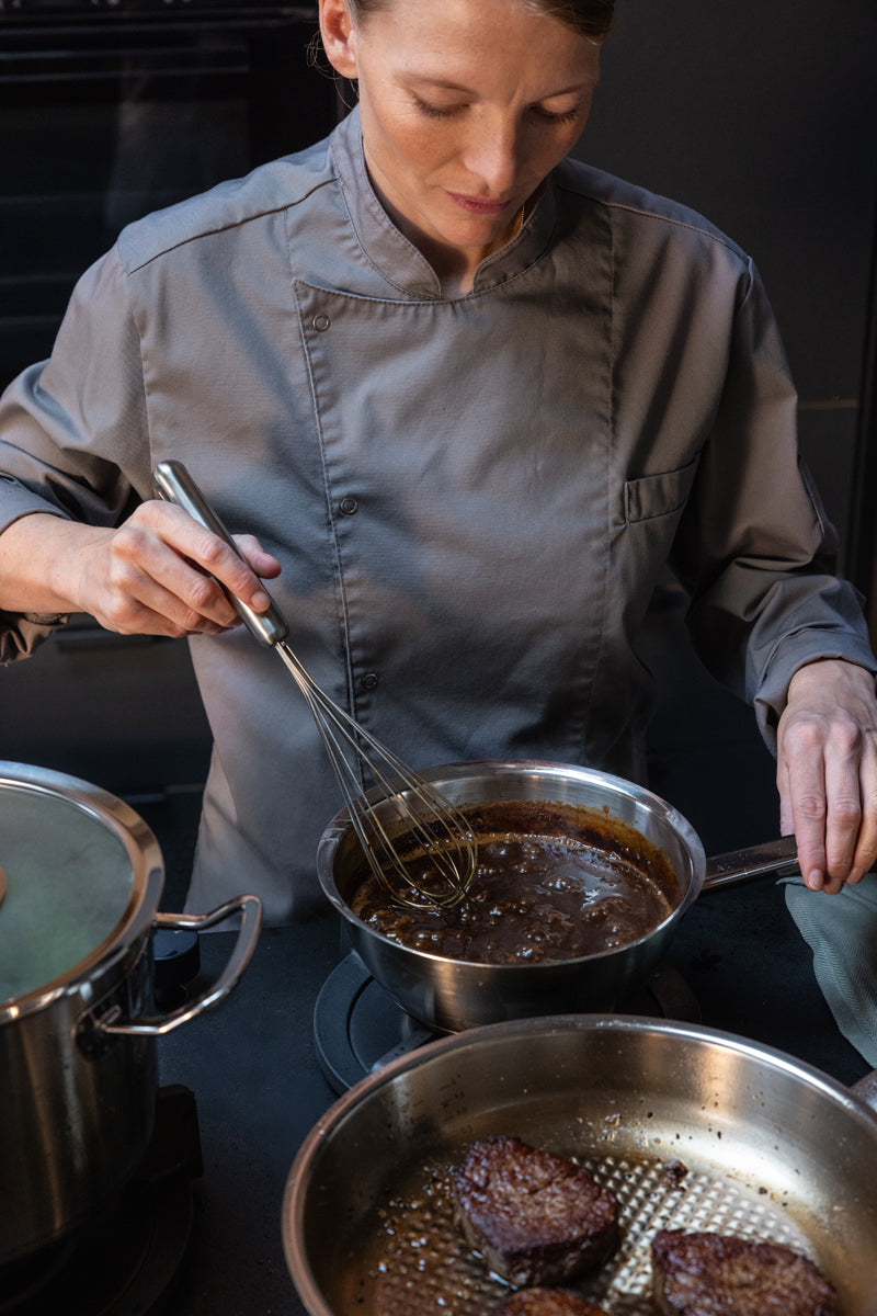 Ein Koch bereitet eine Sauce in einer Fissler Original-Profi Collection® Sauteuse zu; daneben werden in hochwertigen Cookstar-Allherdboden-Pfannen Steaks zubereitet.