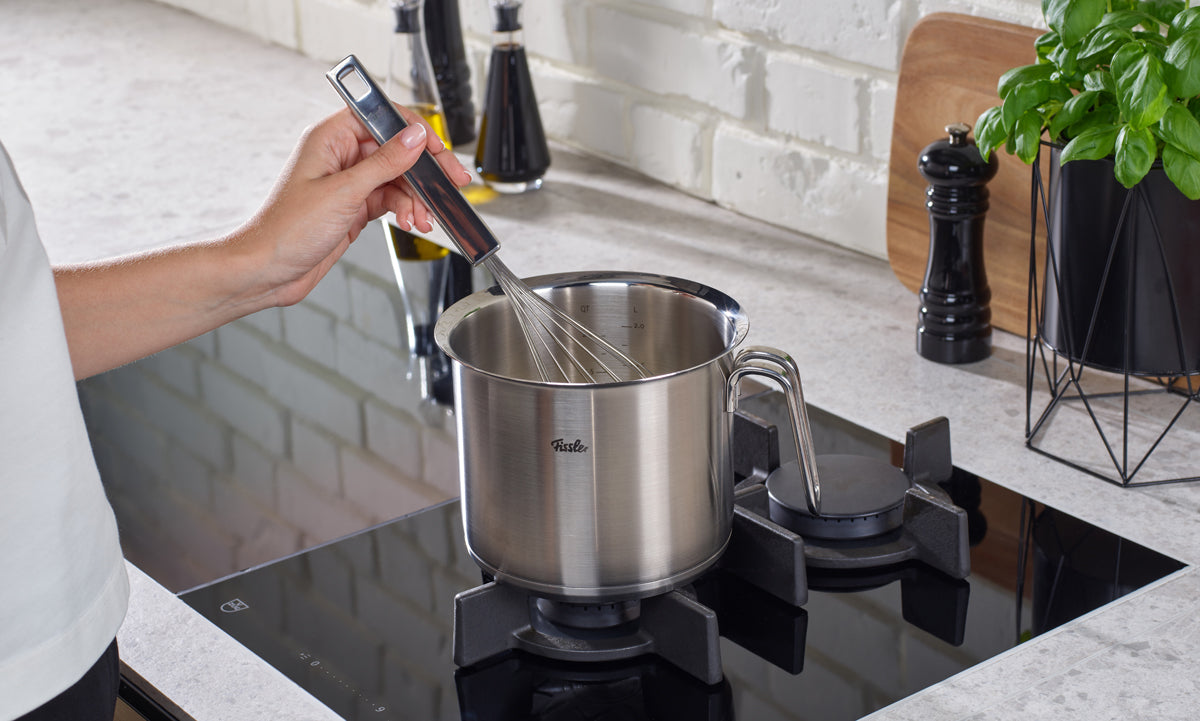Eine Person benutzt einen Metallbesen mit dem Fissler Original-Profi Collection® Milchtopf 16cm auf einem modernen Gasherd in einer hellen Küche.