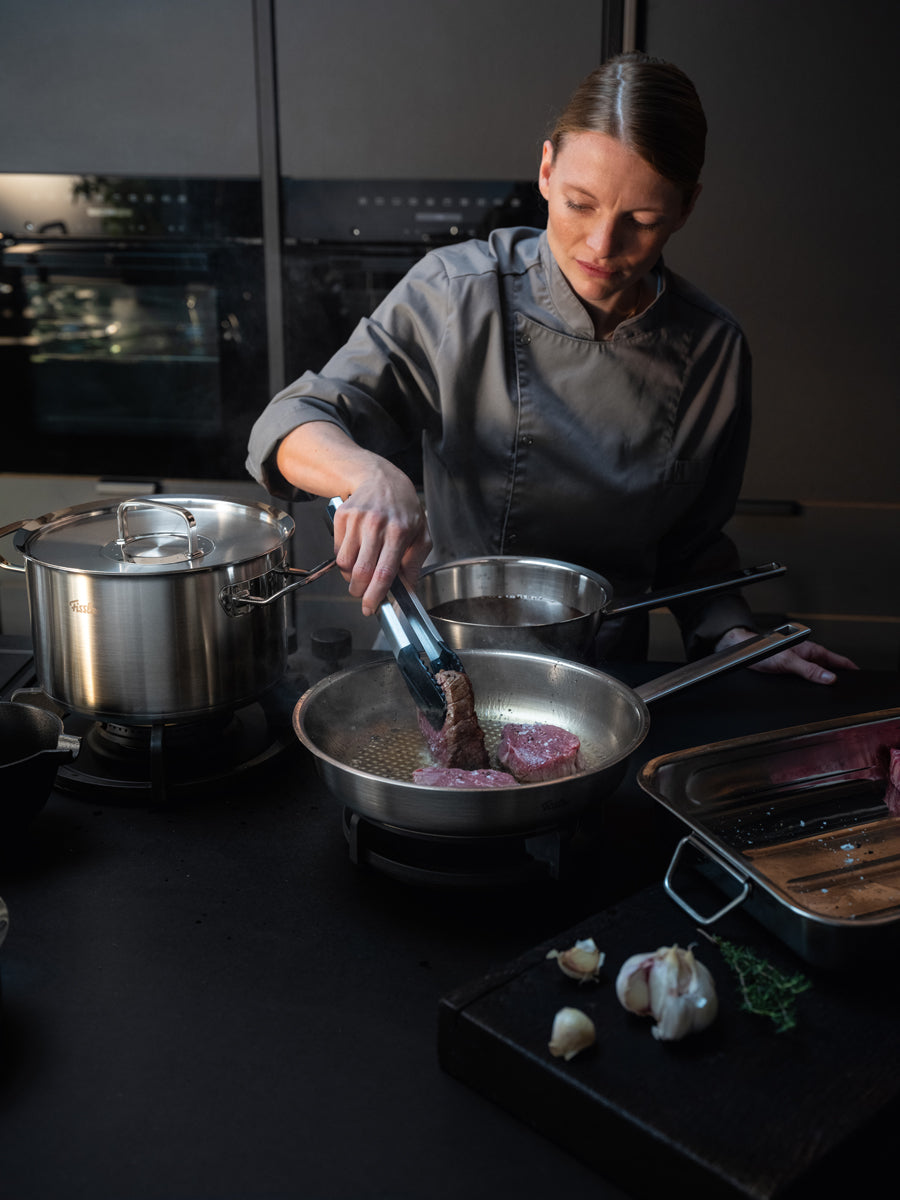 Ein Koch brät Rindfleisch am Herd an; auf dem Tresen stehen Kräuter, Knoblauch und die präzisionsgefertigte Original-Profi Collection® von Fissler.