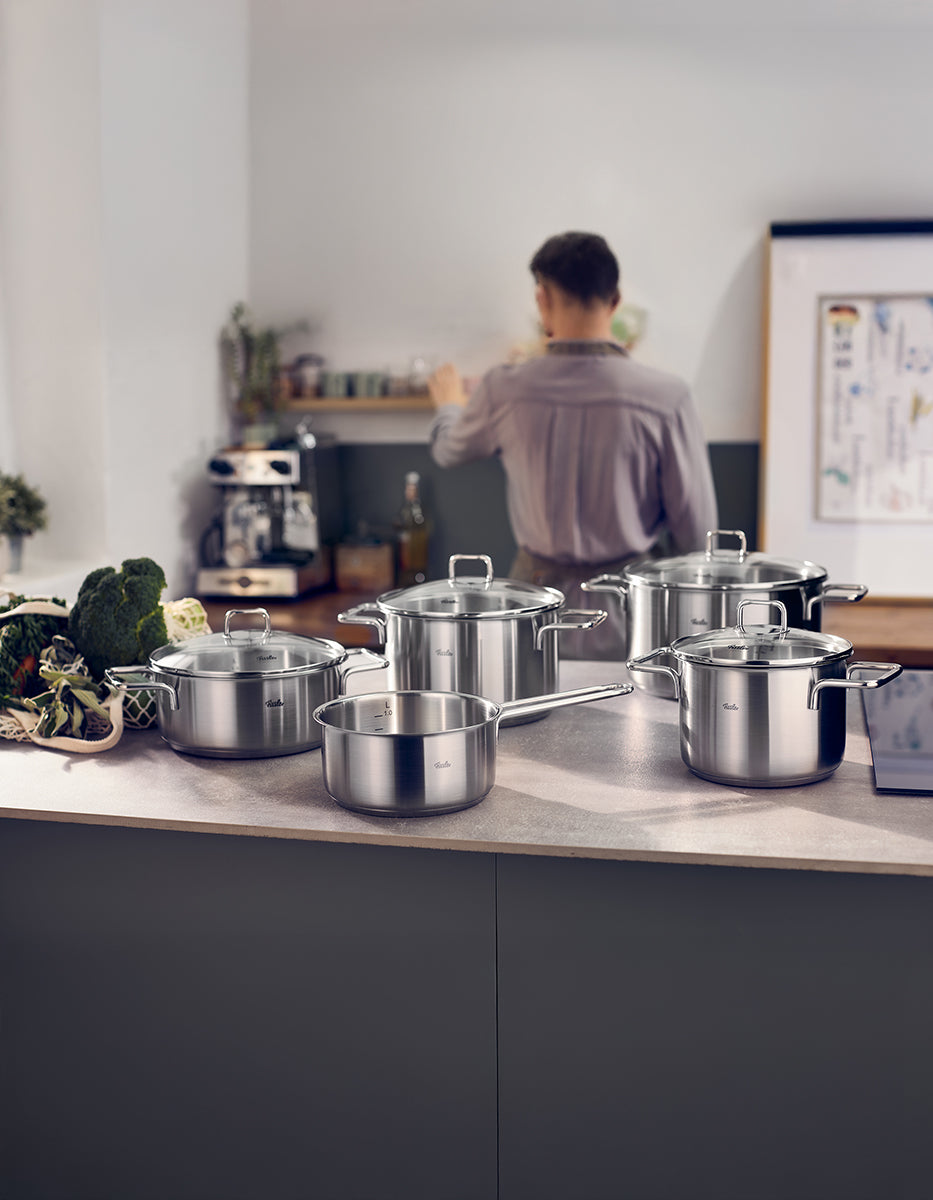Fissler SatiTherm 5-piece stainless steel Cookware Set with glass lids on the counter, suitable for induction; fresh vegetables and shelves in the background.