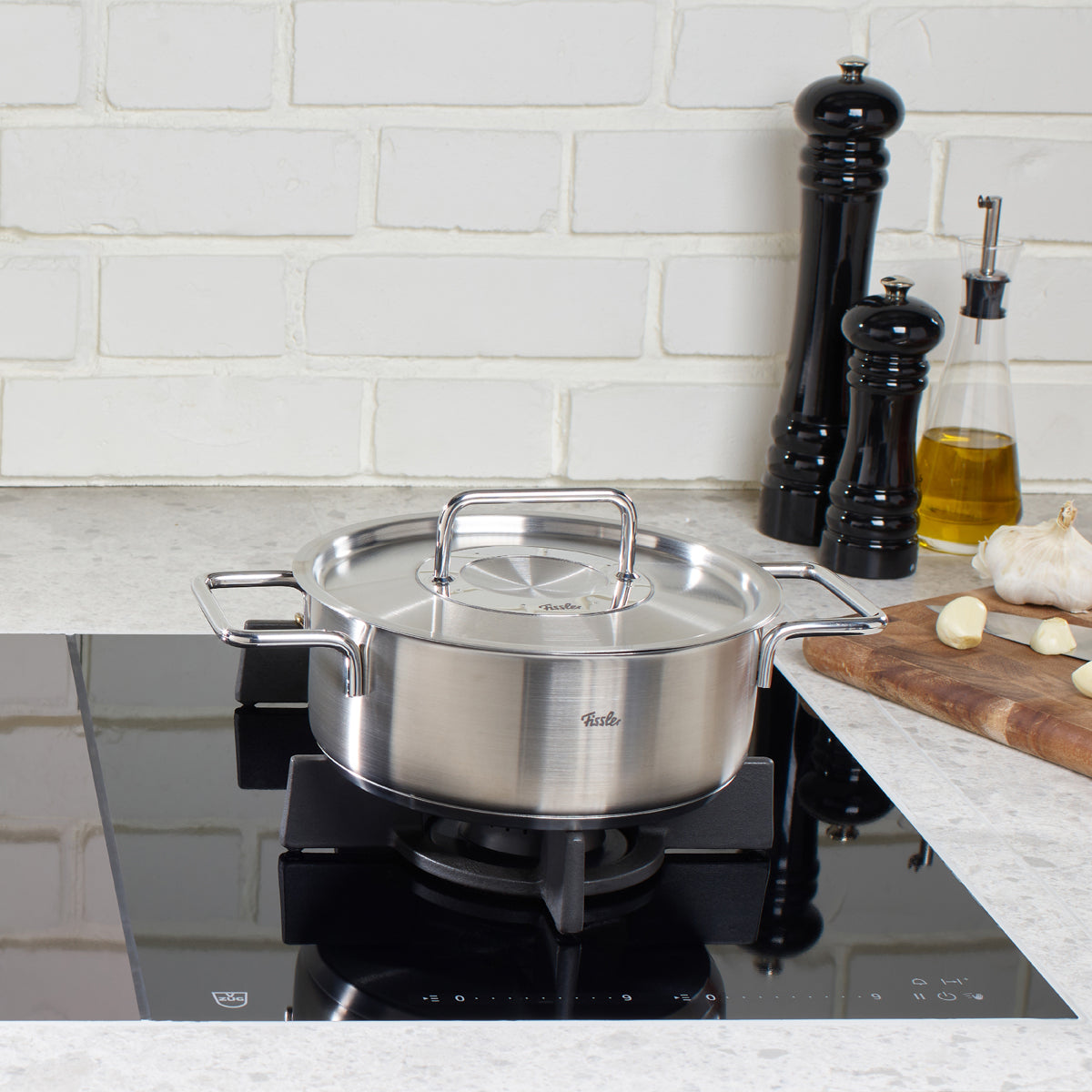 Fissler Pure Collection Casserole Pot with Metal Lid on a black gas stove, surrounded by kitchen utensils and garlic.