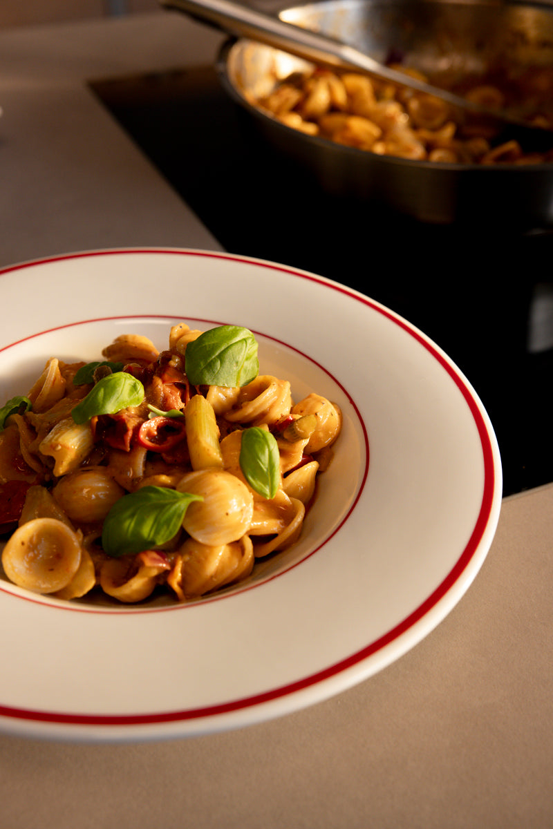 Orecchiette pasta in tomato sauce with basil on a plate; in the background, more pasta in a Fissler Frying Pan on the stove.