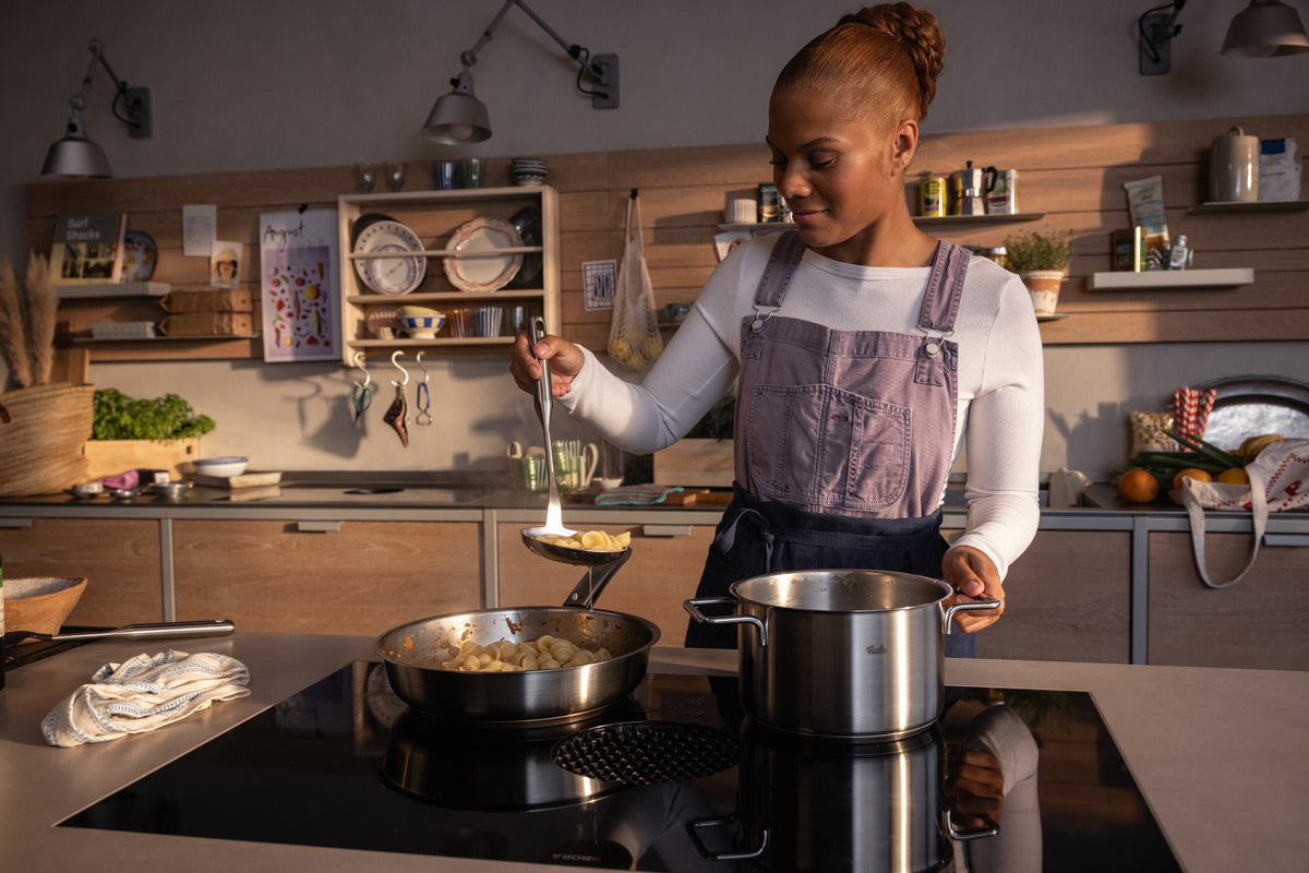 Eine Frau lächelt in einer modernen Küche und kocht mit dem Fissler Pure Collection Kochtopf mit Metalldeckel, bekannt für Qualität Made in Germany.