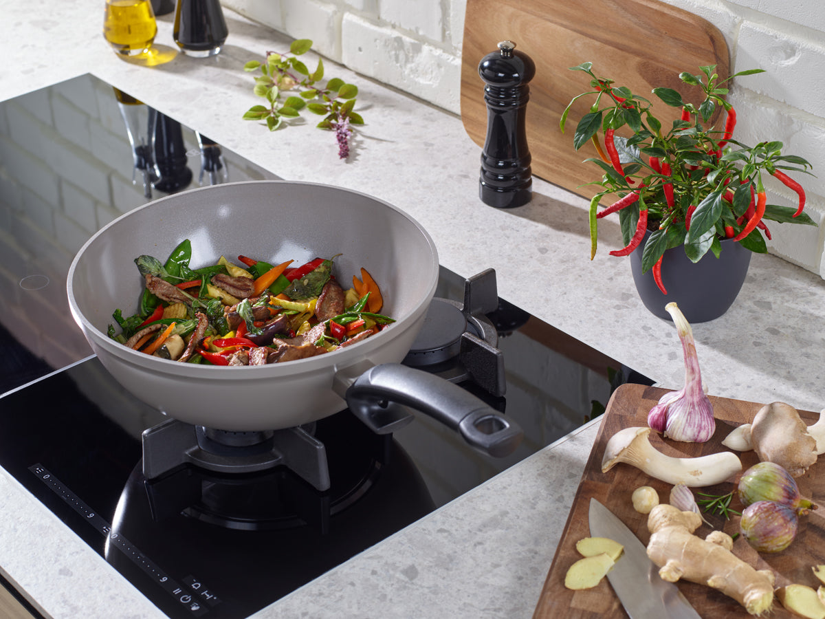 Fissler Ceratal® Wokpfanne mit Rührbraten auf dem Herd, Kräutern, Pfeffermühle, Chili, Knoblauch & Ingwer - hochwertiges deutsches Kochgeschirr.